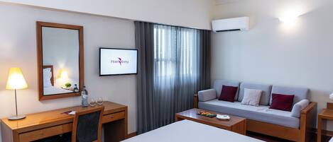 Une chambre d’hôtel comprenant un lit, un bureau, une chaise, un canapé, une table basse, une lampe, un miroir, un cadre photo, une fenêtre avec des rideaux et un appareil de climatisation.