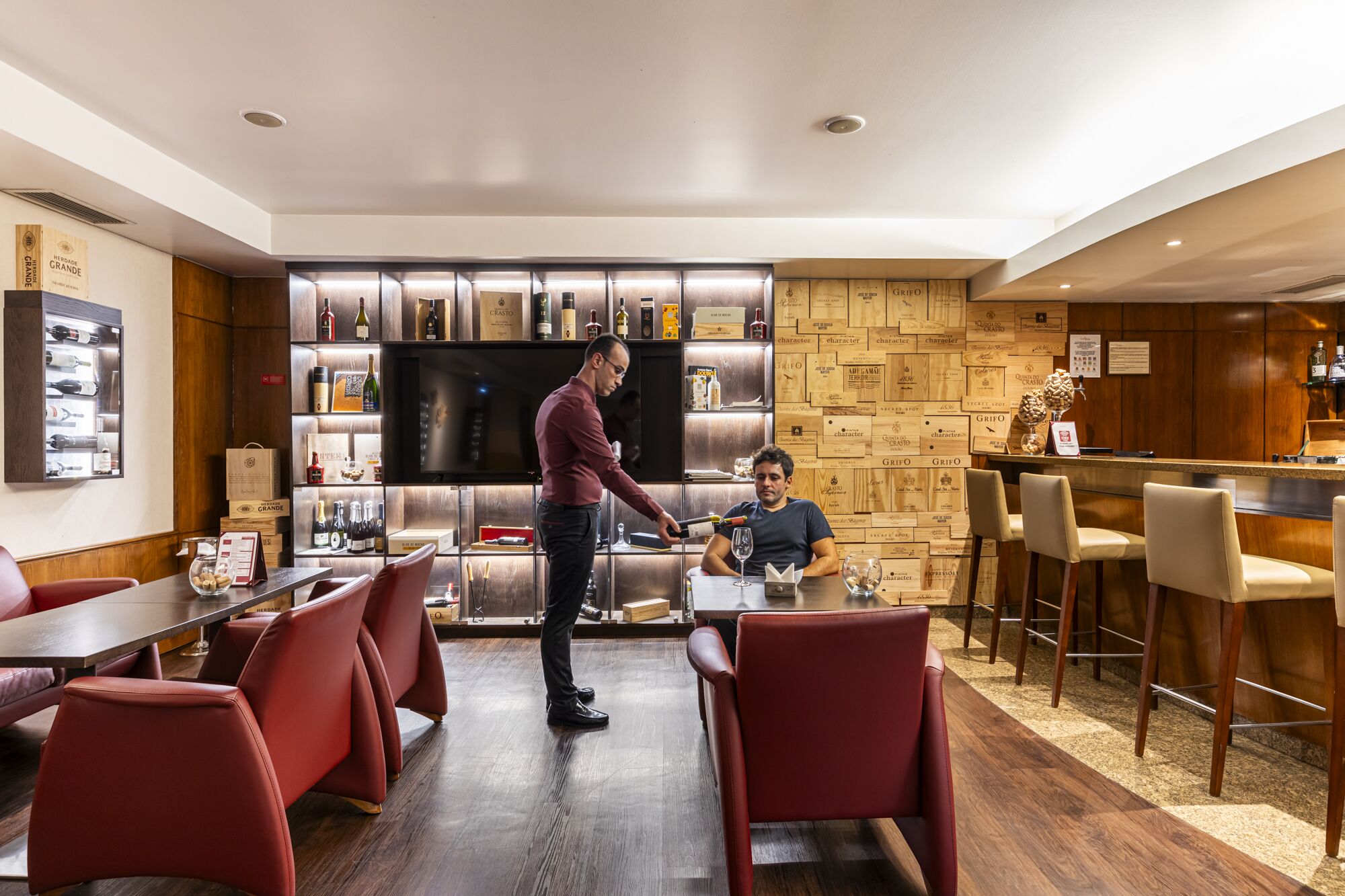 A restaurant with a bar area, wooden wall decor, and red chairs.