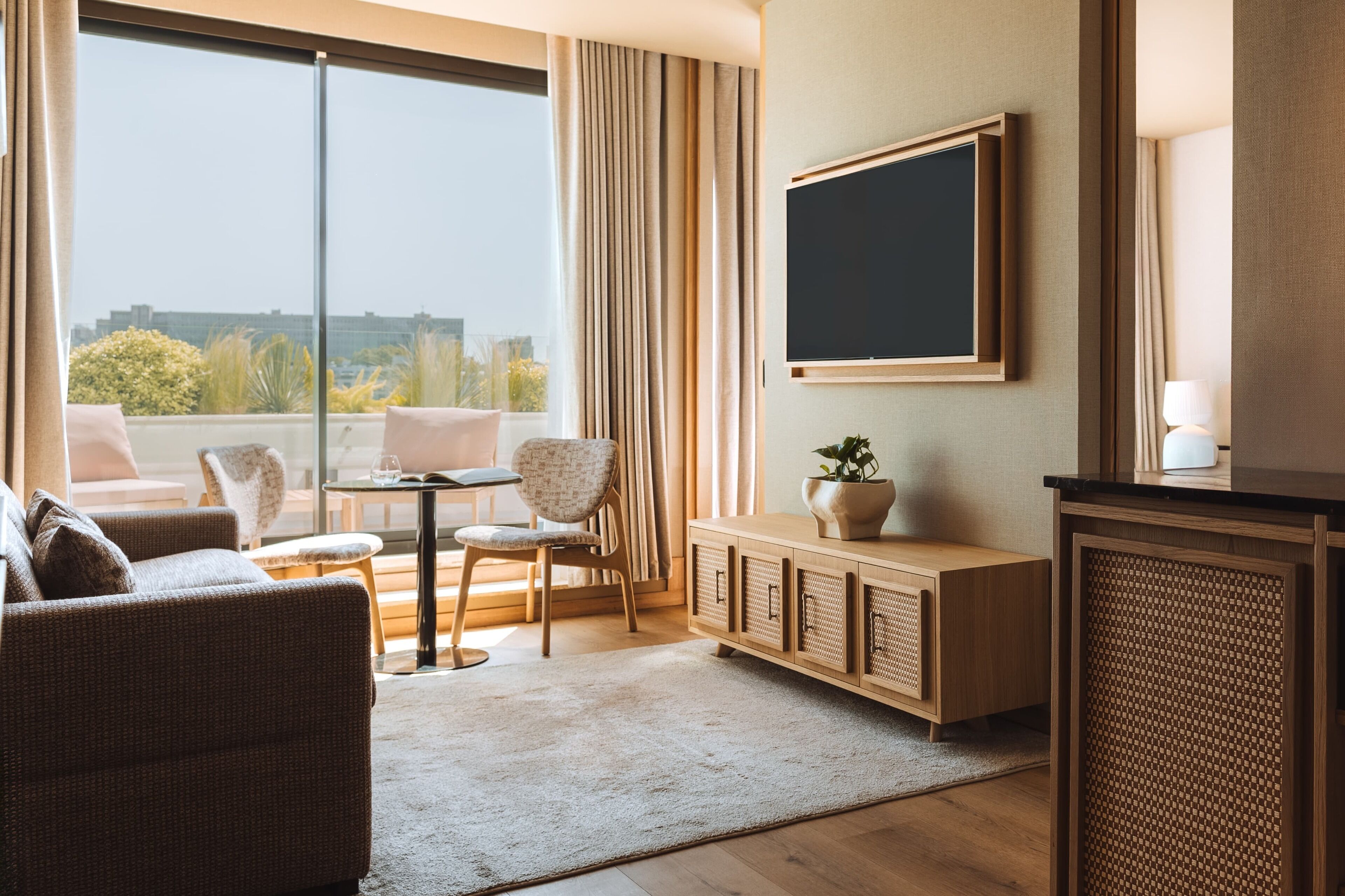 A modern hotel room with a large window, a flat-screen TV, a sofa, a dining table, and a cabinet.