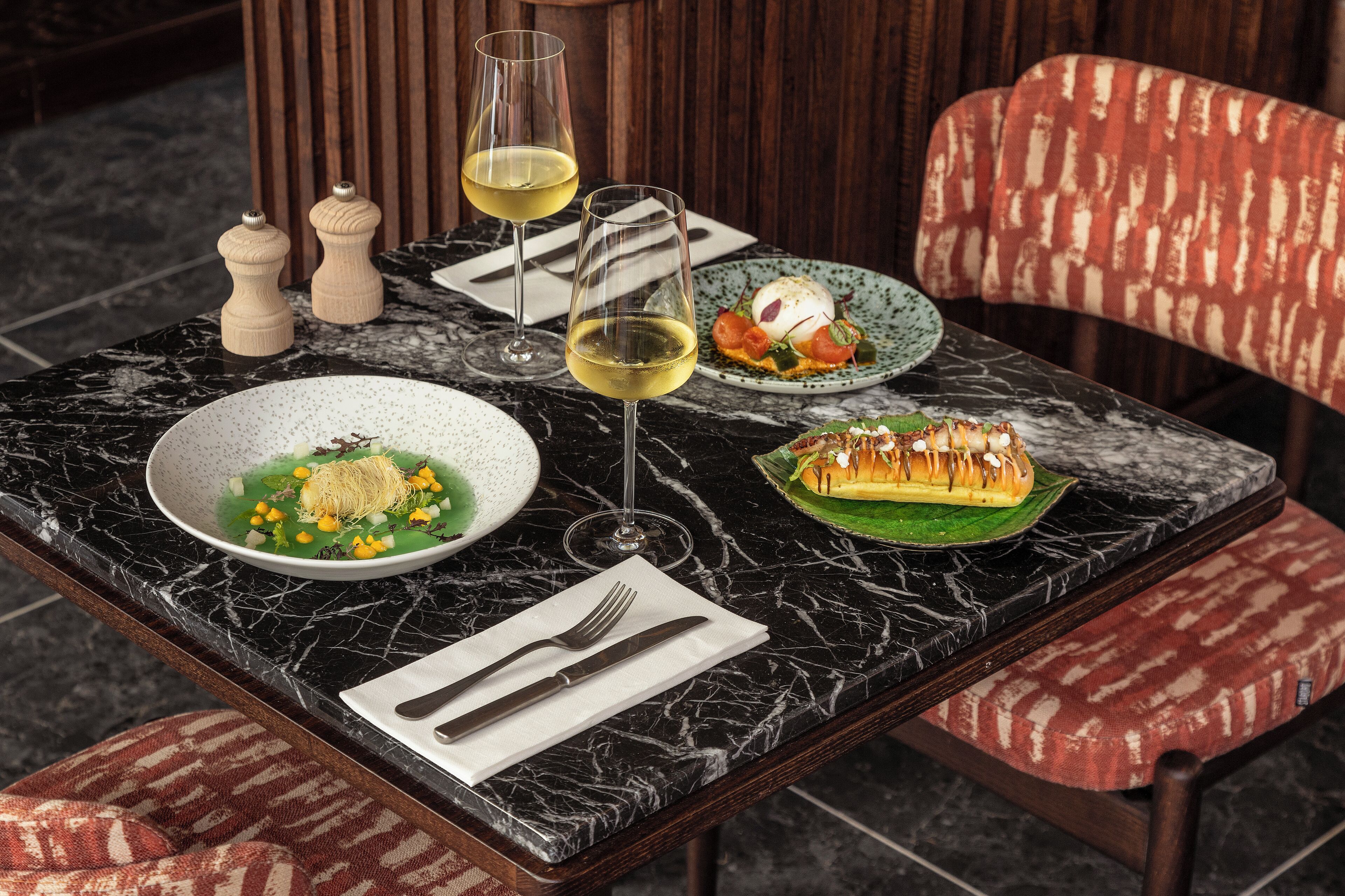 Una mesa de comedor con dos copas de vino, un tazón de sopa, un plato de verduras y un postre sobre una encimera de mármol.