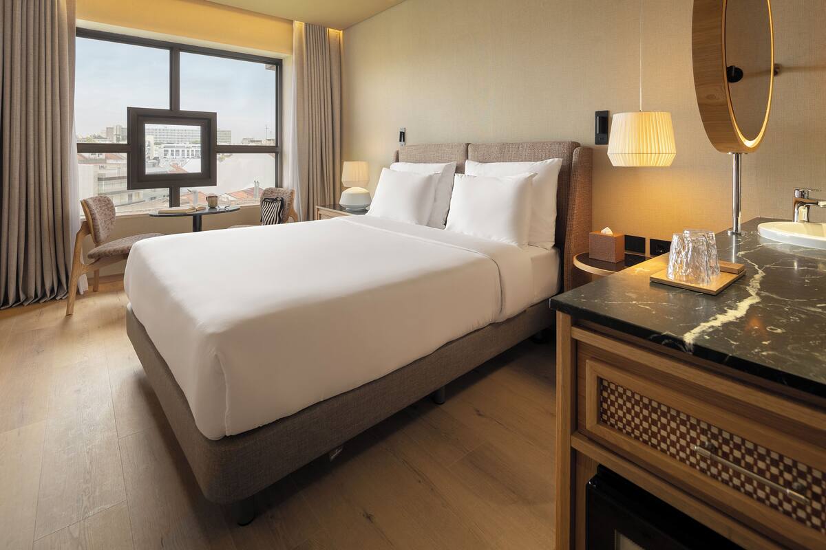 A hotel room with a large bed, a desk with a computer, a chair, a window with curtains, and a bathroom with a mirror and sink.