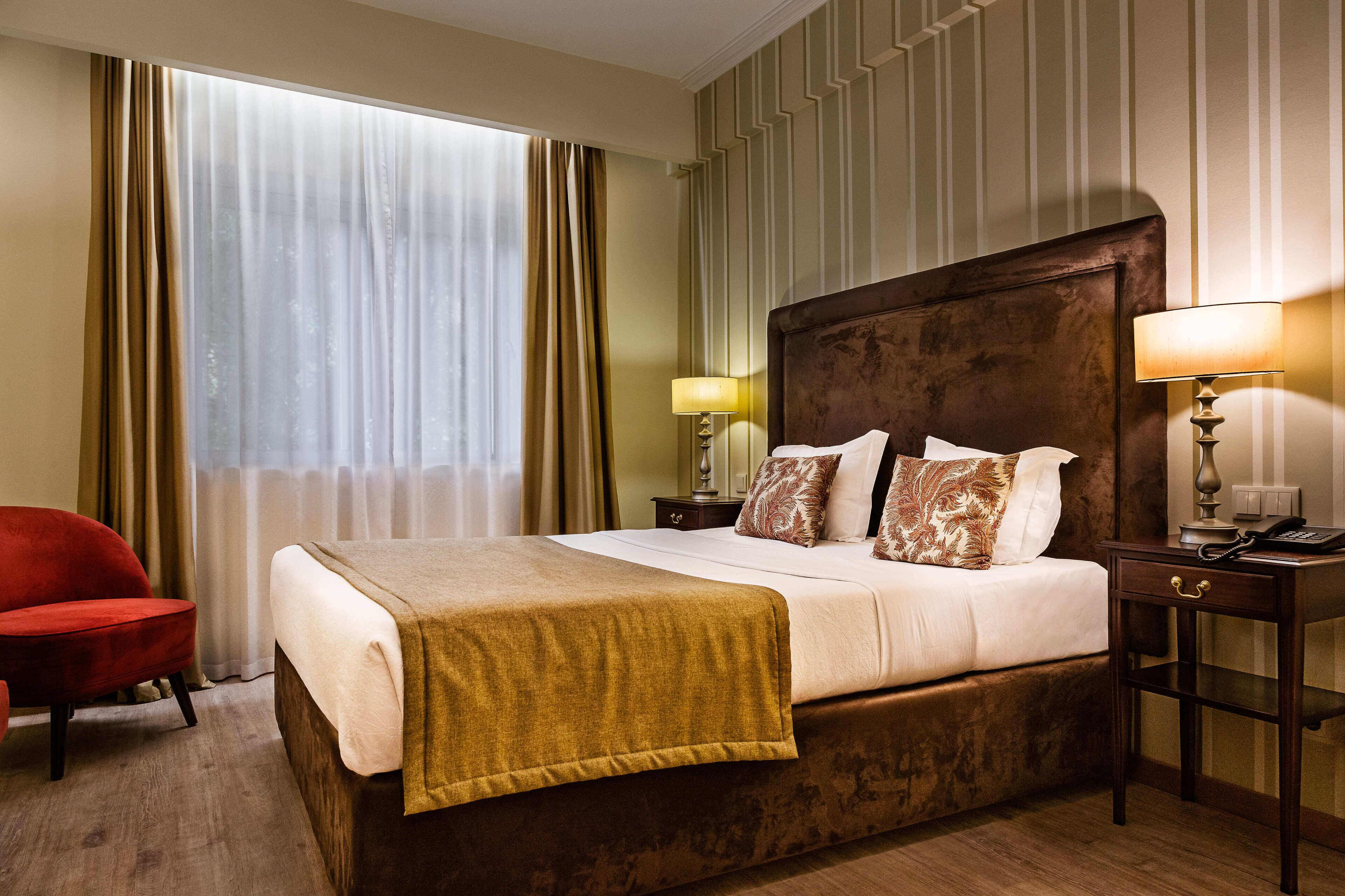 A hotel room with a large bed, a bedside table with a lamp, a red chair, and a window with curtains.