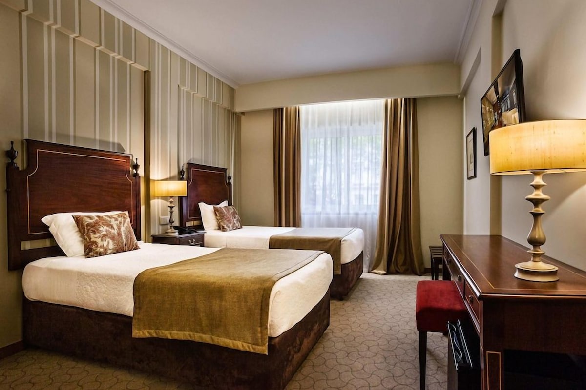A hotel room with two beds, a desk, a chair, a lamp, and a window with curtains.