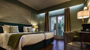 A hotel room with a large bed, a desk, and a chair. There is a window with curtains and a view of trees.