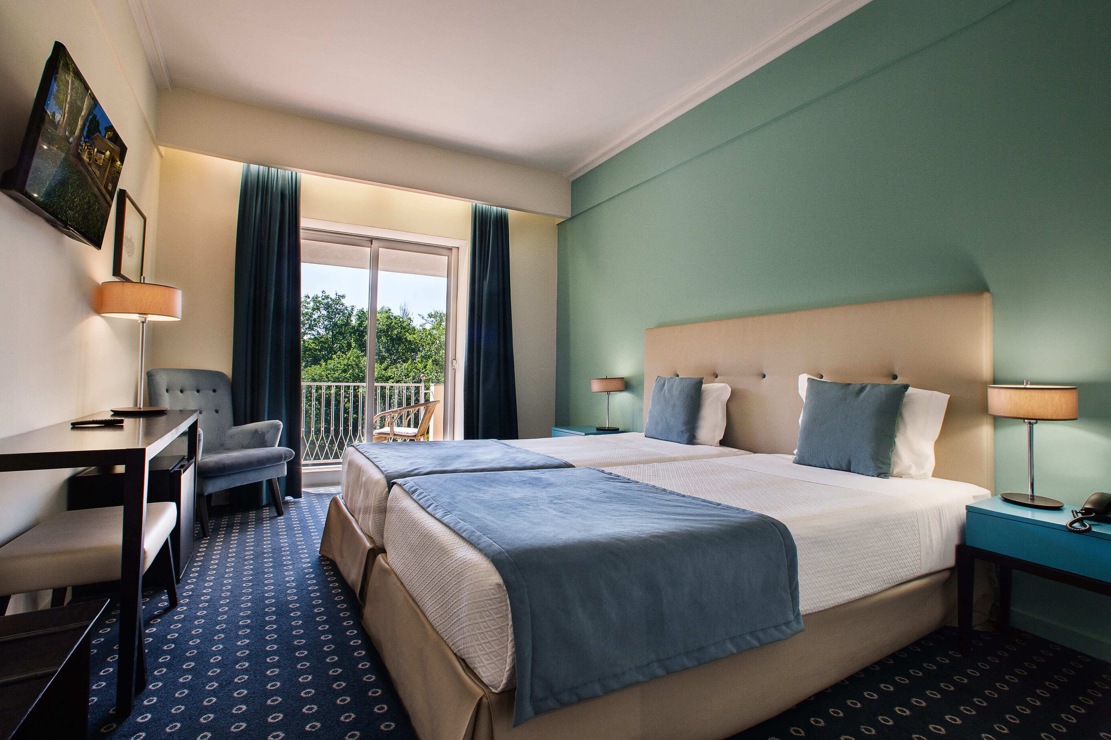 A hotel room with two beds, a desk, a chair, a balcony with a view, and a painting on the wall.