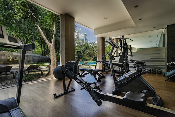 A modern gym with various exercise equipment, large windows, and a view of a swimming pool.