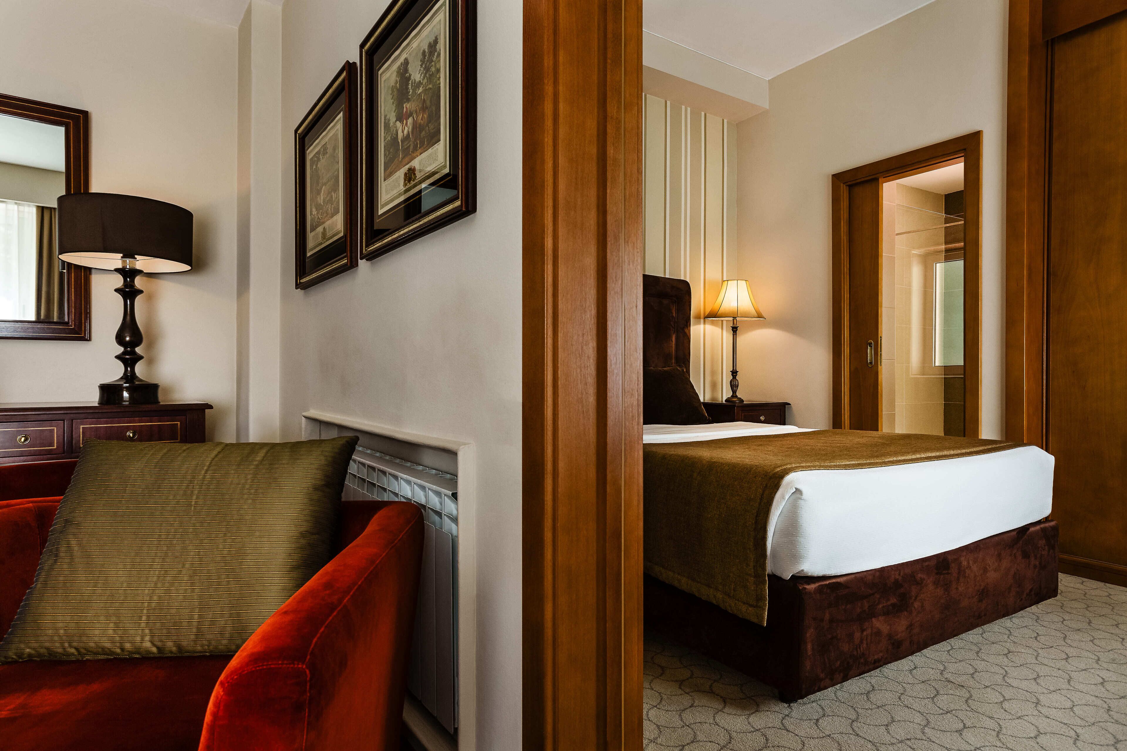 Habitación de hotel con cama, un sillón rojo, una mesita de noche con lámpara y cuadros en la pared.