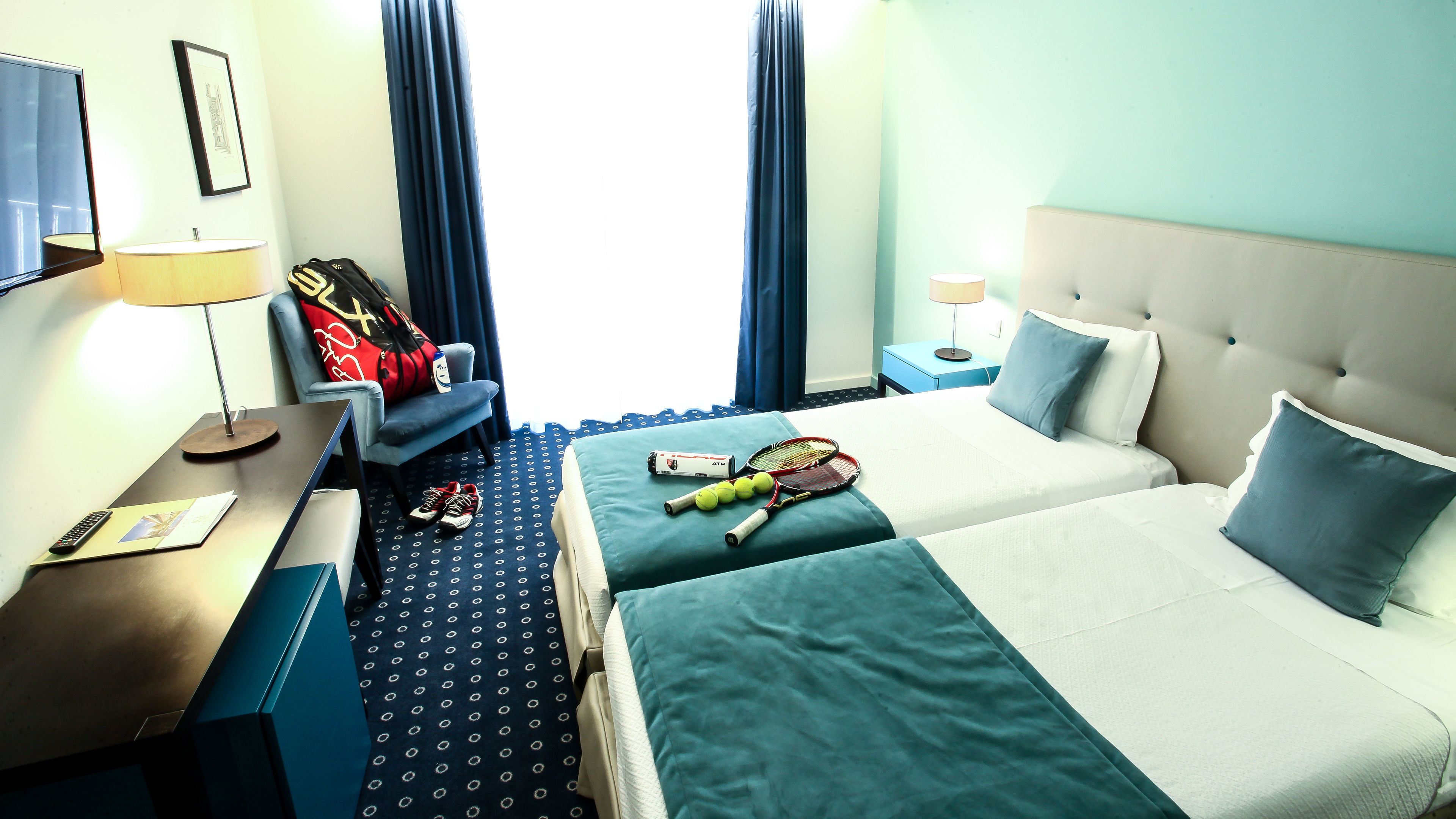 A hotel room with a bed, a desk, a chair, a lamp, and a window with curtains.