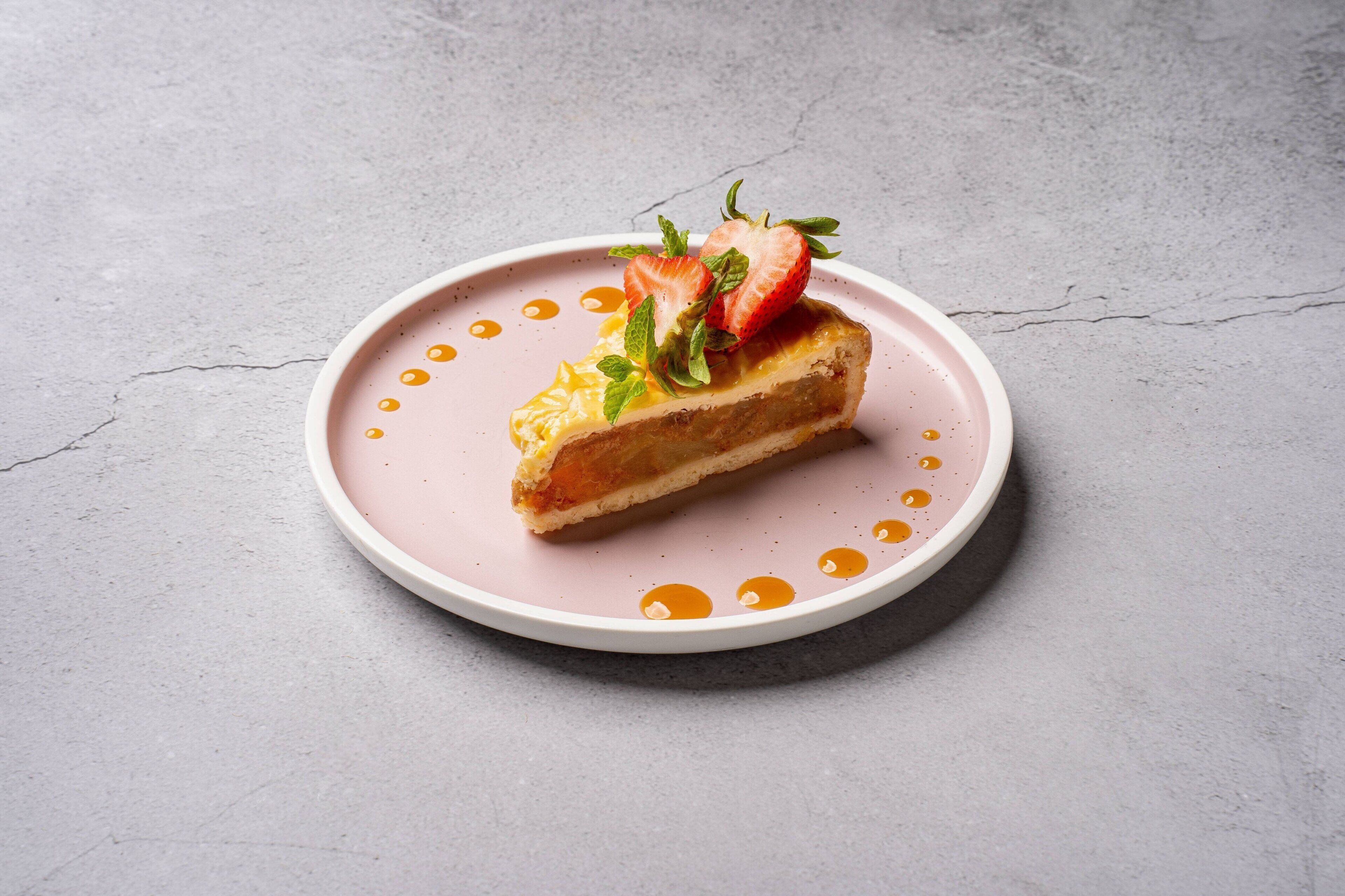 Ein Stück Kuchen auf einem rosafarbenen Teller, mit Sirup und Erdbeeren auf einer grauen Unterlage.