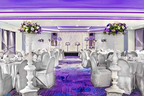 Une salle de banquet avec des rangées de tables, des chaises blanches et des centres de table floraux violets.
