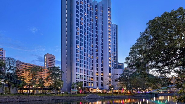 Un hotel moderno de gran altura con varios pisos, un patio cuidado y una masa de agua que refleja el edificio.