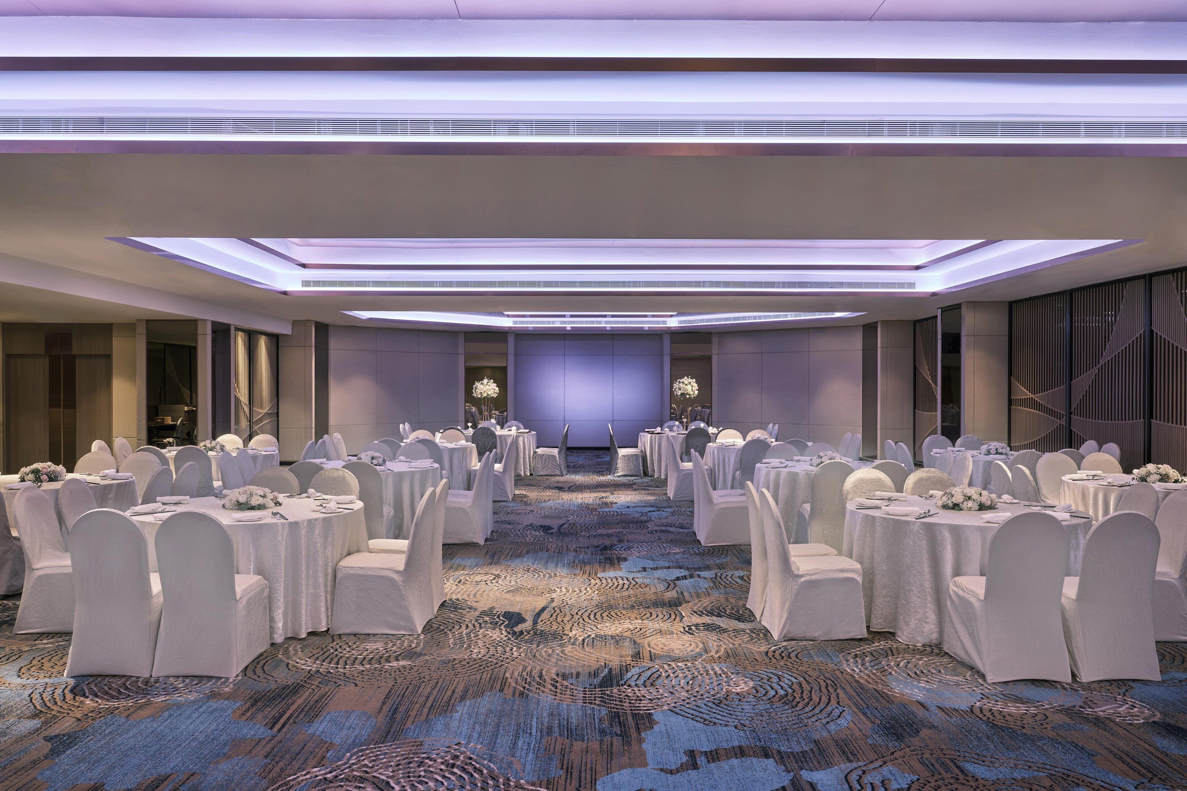 Un salón de banquetes con mesas redondas y sillas blancas, una alfombra estampada y un escenario al frente.