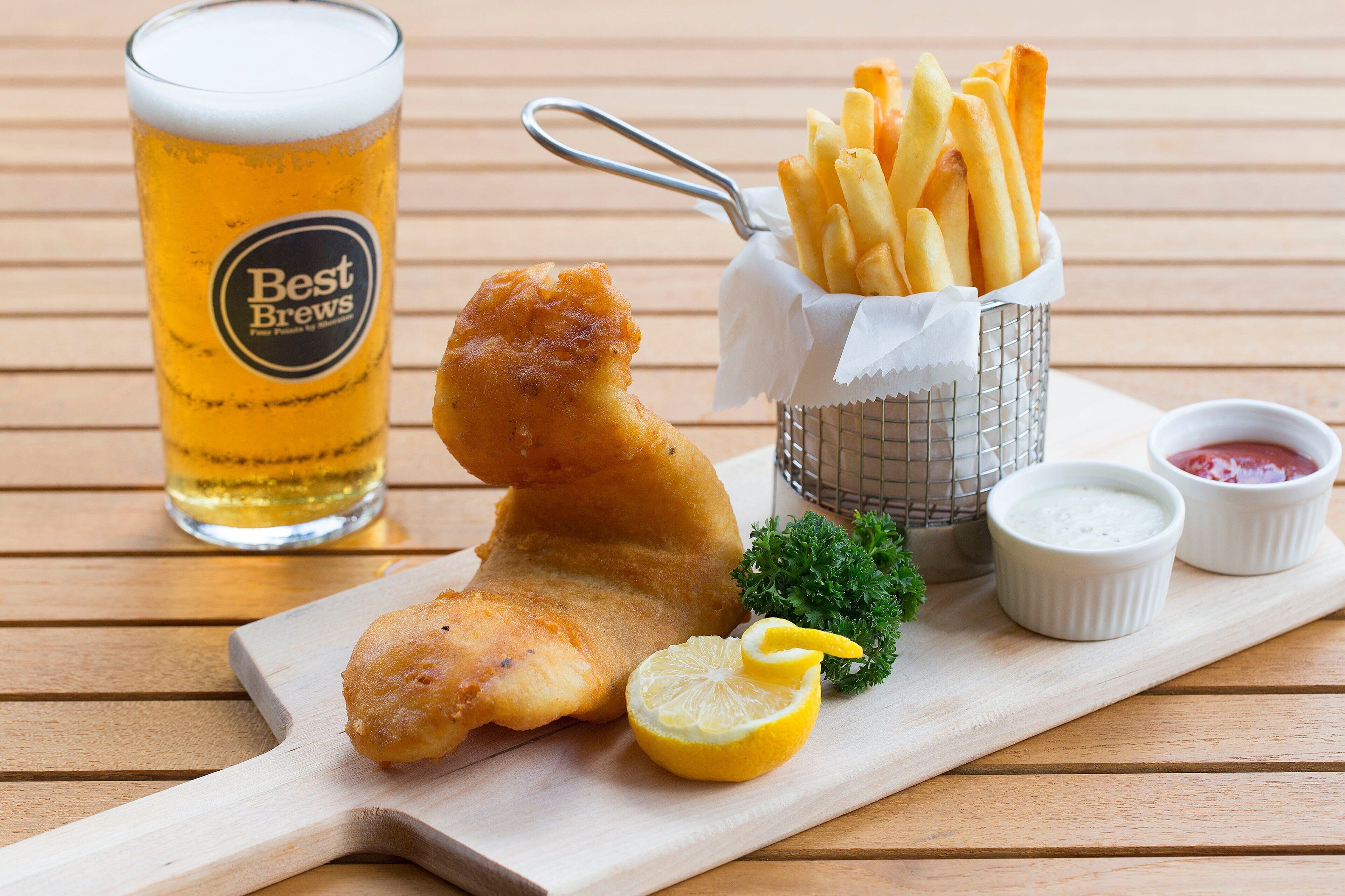 Un plato de pescado con papas fritas, una cerveza en vaso y dos salsas pequeñas para mojar, todo sobre una mesa de madera.