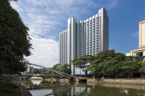 Un hôtel moderne de grande hauteur, avec une rivière au premier plan, un pont piétonnier et une végétation luxuriante.