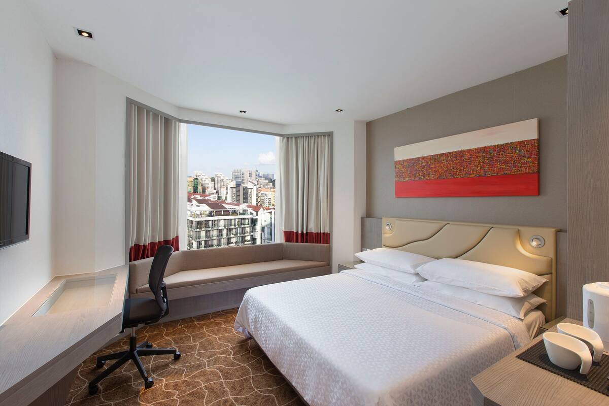 Une chambre d’hôtel avec un grand lit, un bureau avec une chaise, une fenêtre avec des rideaux, une vue sur la ville et un tableau au mur.