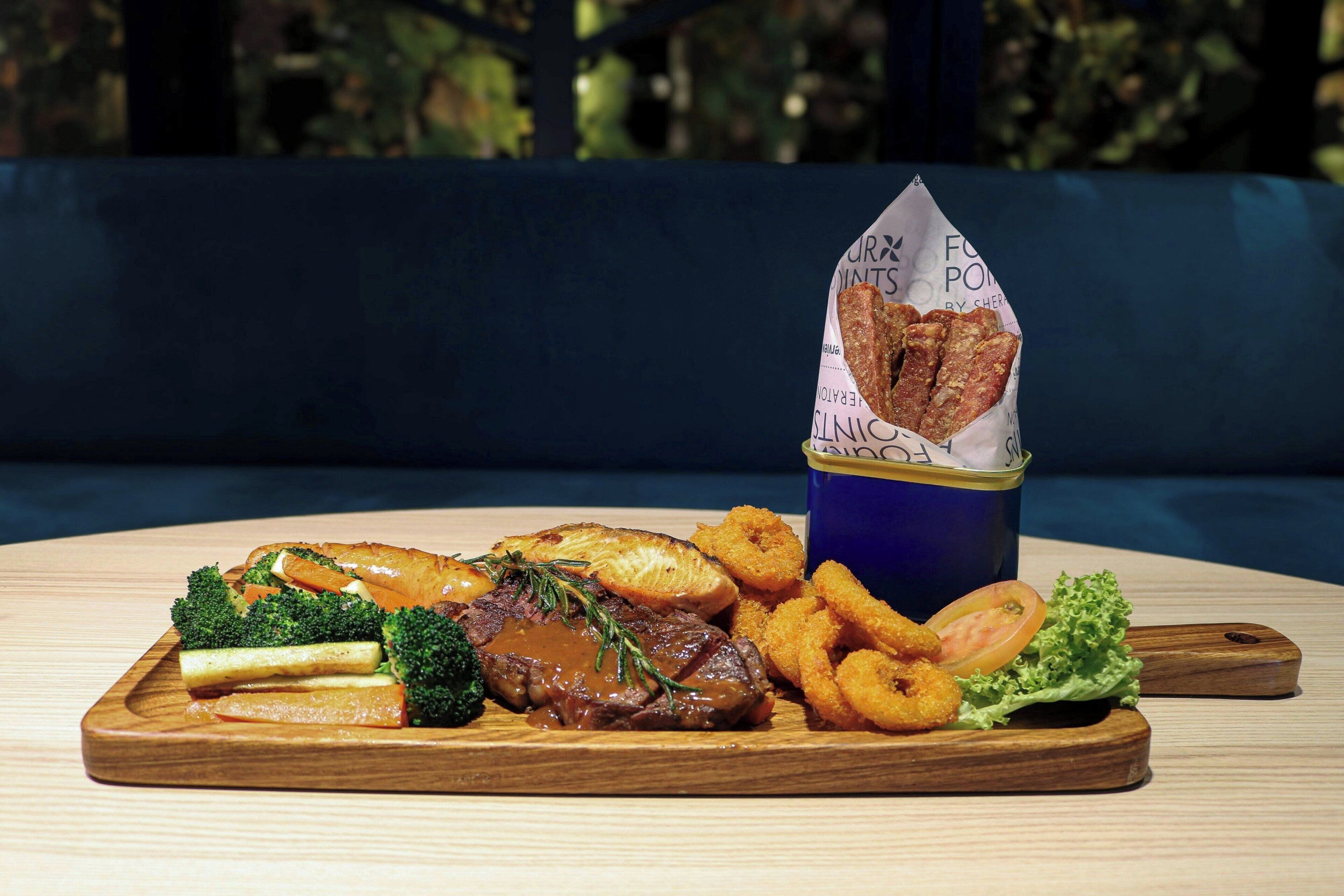 Une planche de service en bois, sur laquelle sont disposés divers aliments, notamment de la viande, des légumes et des plats frits, accompagnée d’une tasse bleue avec une étiquette.