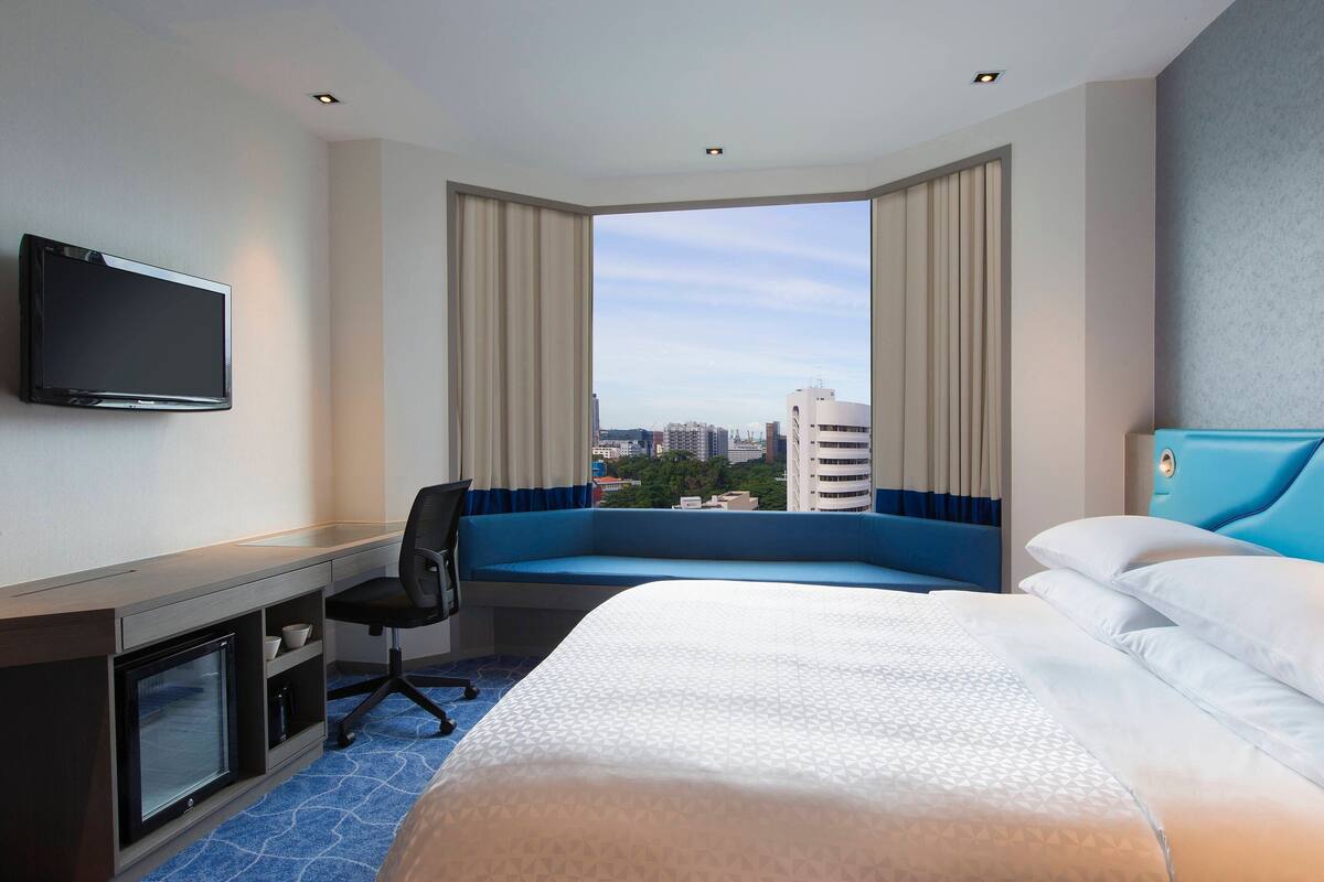 Une chambre d’hôtel avec un lit, un bureau équipé d’un ordinateur, une télévision et une grande fenêtre avec des rideaux.