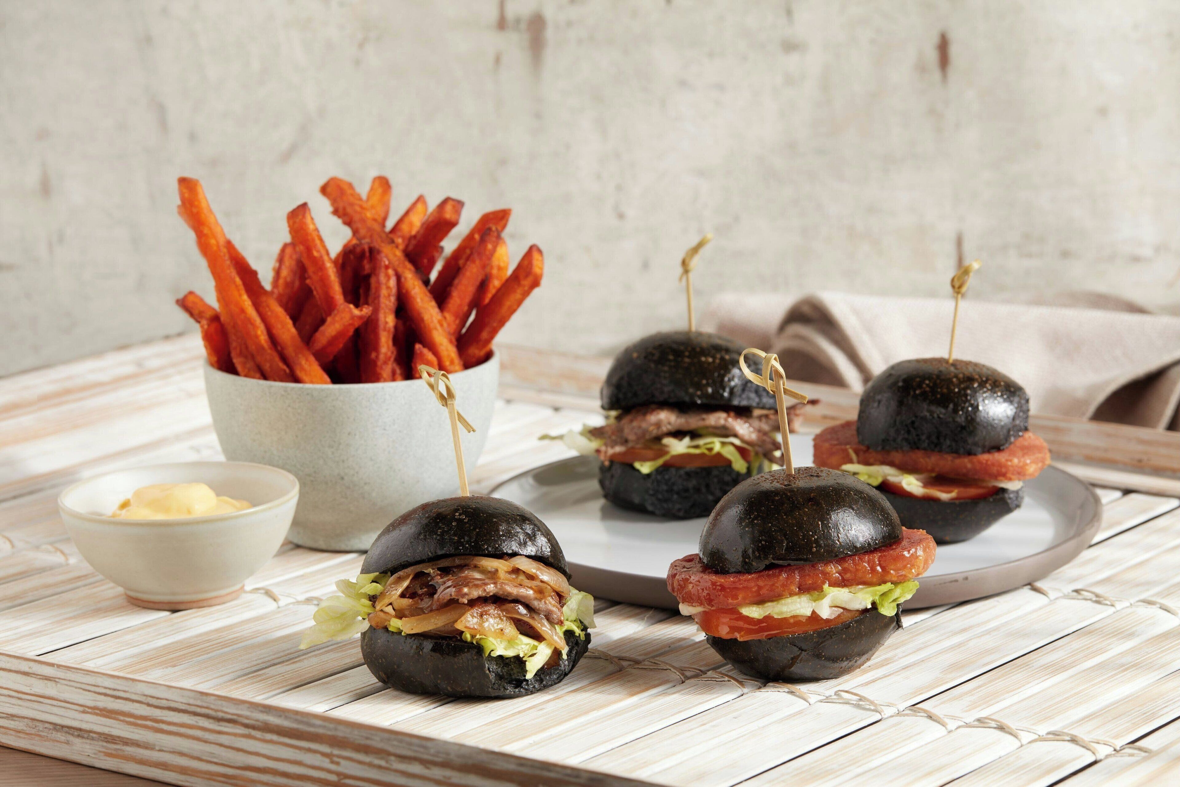 Three burgers with black buns and toothpicks, a bowl of fries, and a small bowl of butter on a wooden table.