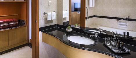 A modern bathroom with a black countertop, a white sink, and a mirror. There are towels on a shelf and a red suitcase on the floor.