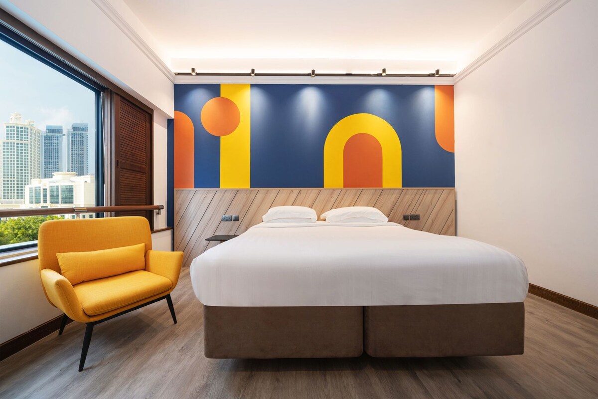 A hotel room with a large bed, a yellow armchair, a city view, and a colorful abstract wall mural.