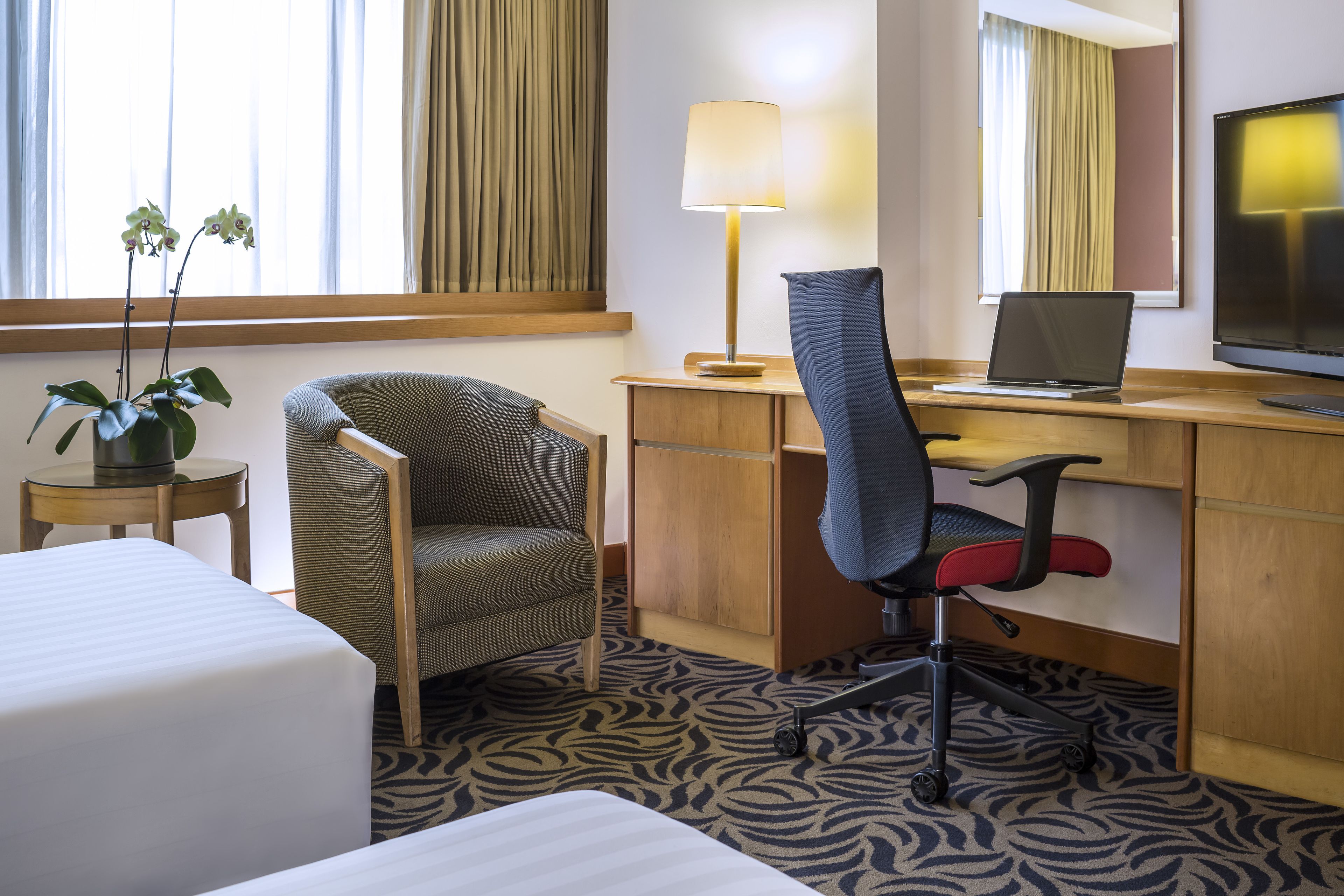 A hotel room with a bed, a desk with a laptop, a chair, a lamp, and a TV.
