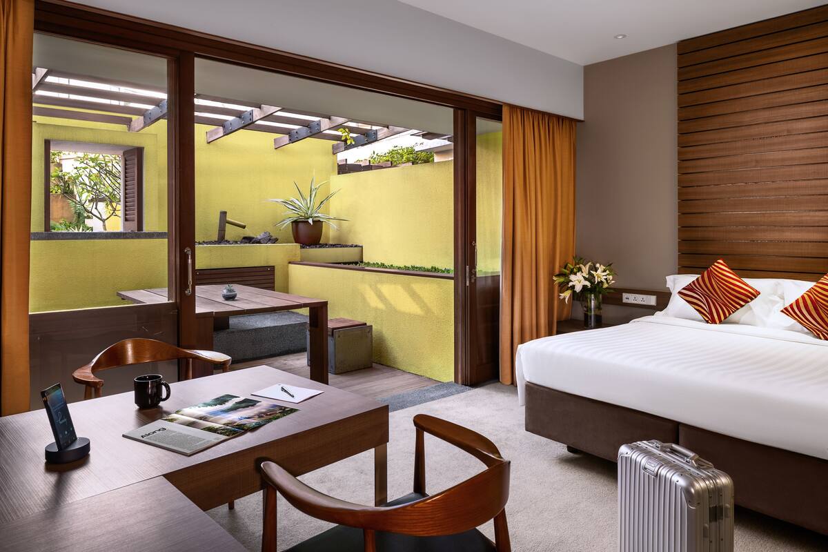 A hotel room with a bed, a desk with a chair, a small table, a potted plant, and a suitcase.