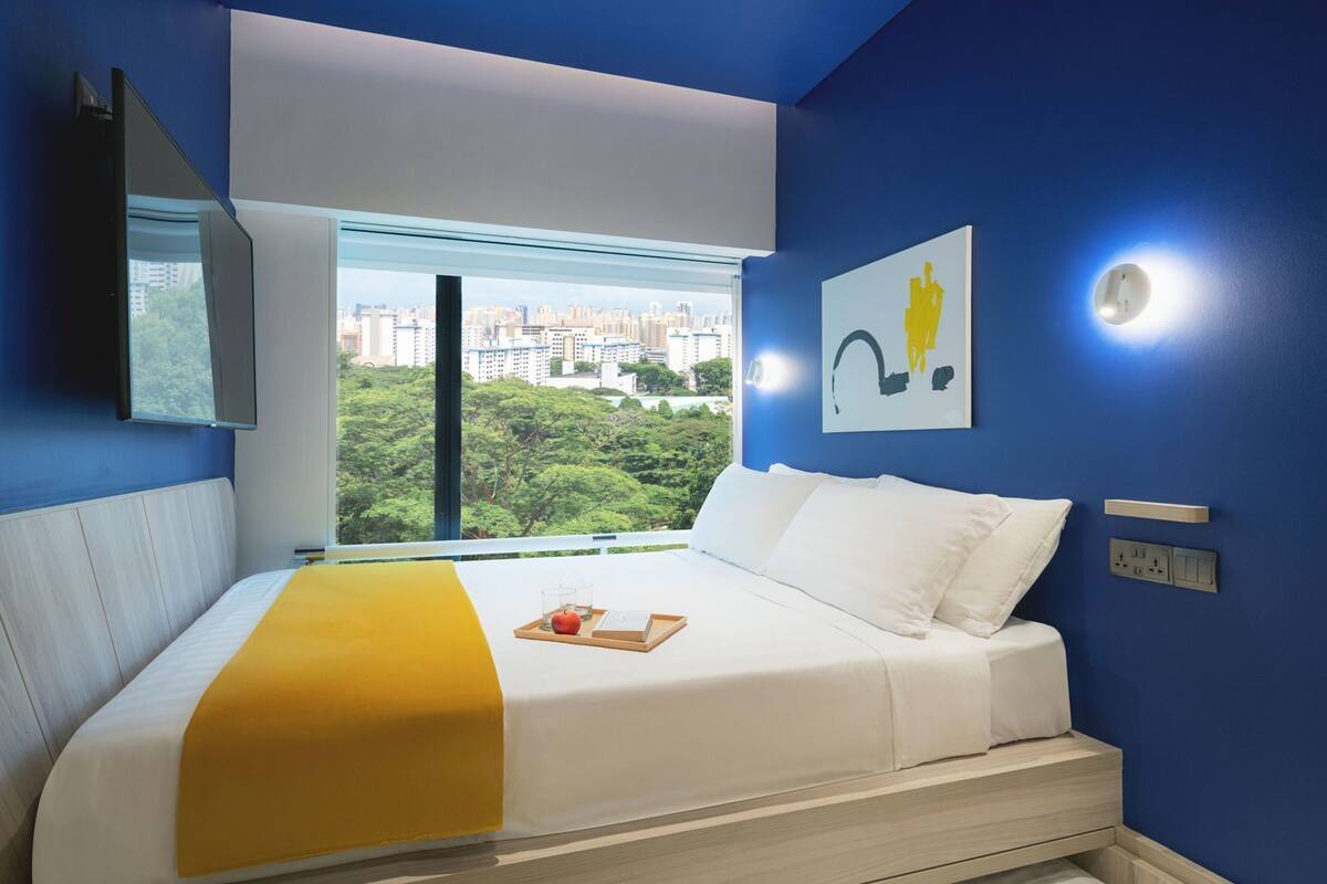 A hotel room with a bed, a window with a city view, a mounted TV, and a painting on the wall.