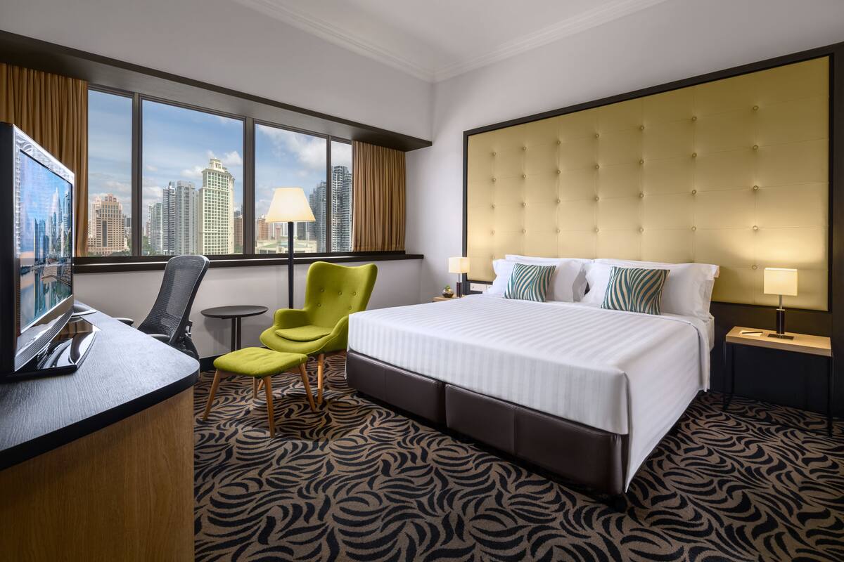 A hotel room with a large bed, a desk with a chair, a small table, and a view of the cityscape through large windows.