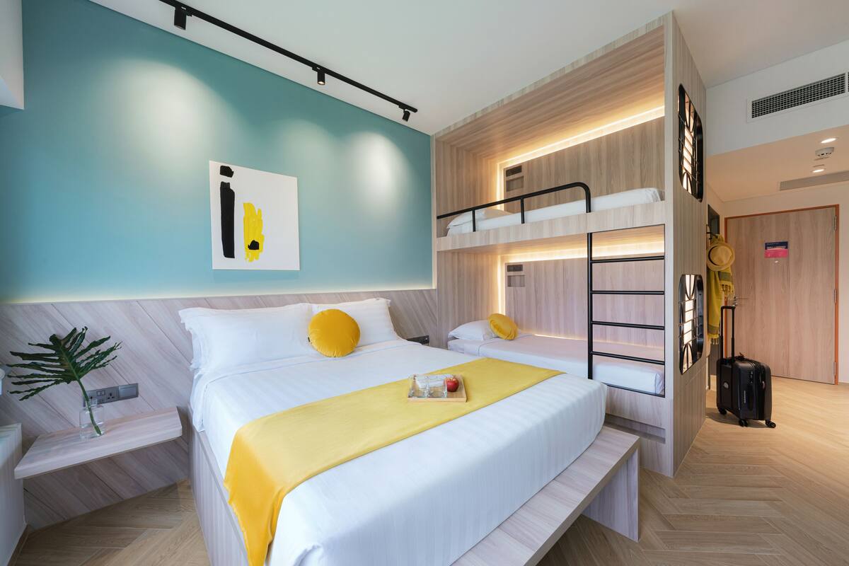 A modern hotel room with a bunk bed, a bedside table with a plant, a yellow bedspread, and a suitcase in the corner.