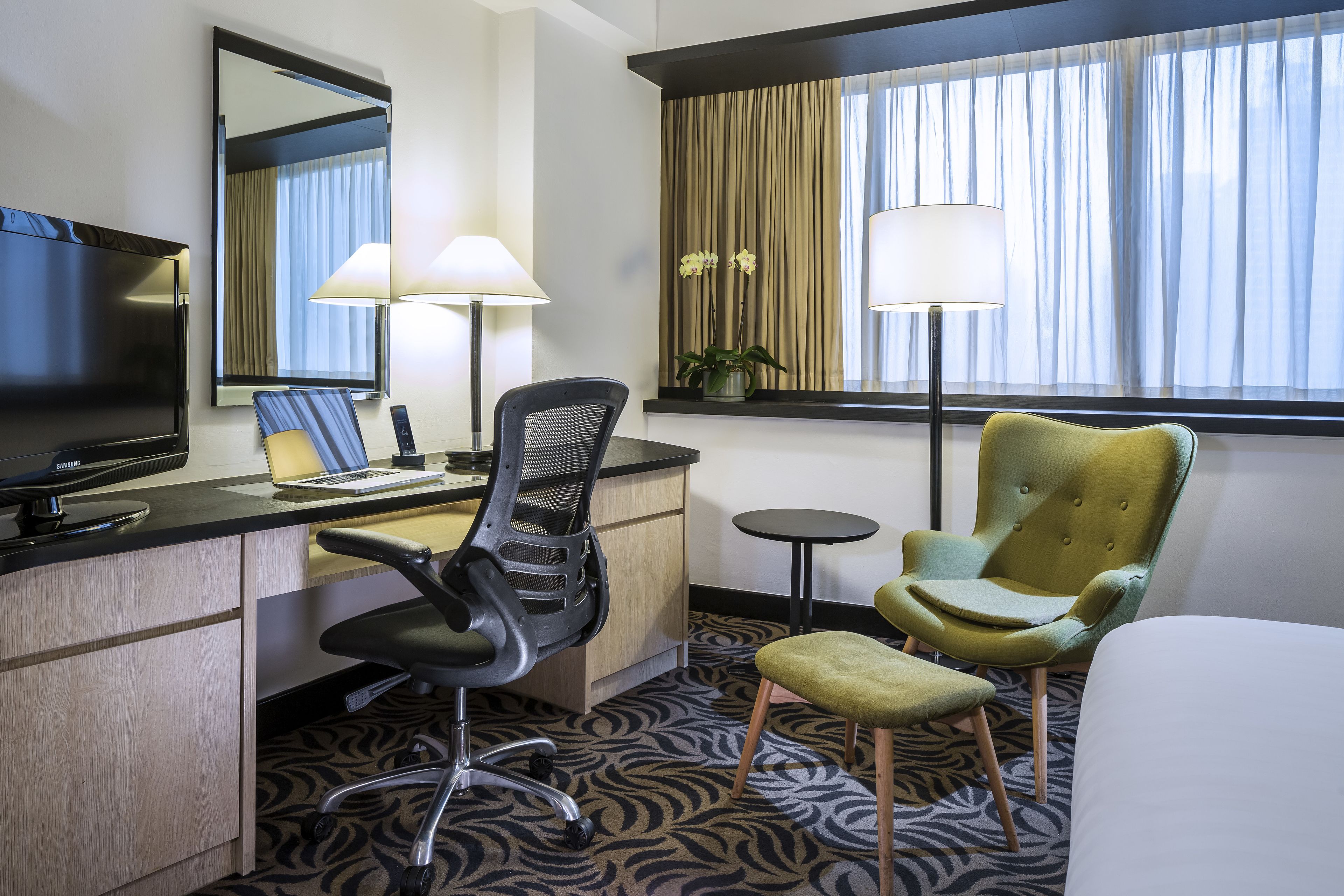 A hotel room with a flat-screen TV, a desk with a laptop, a chair, a small table, a lamp, a potted plant, and a bed.