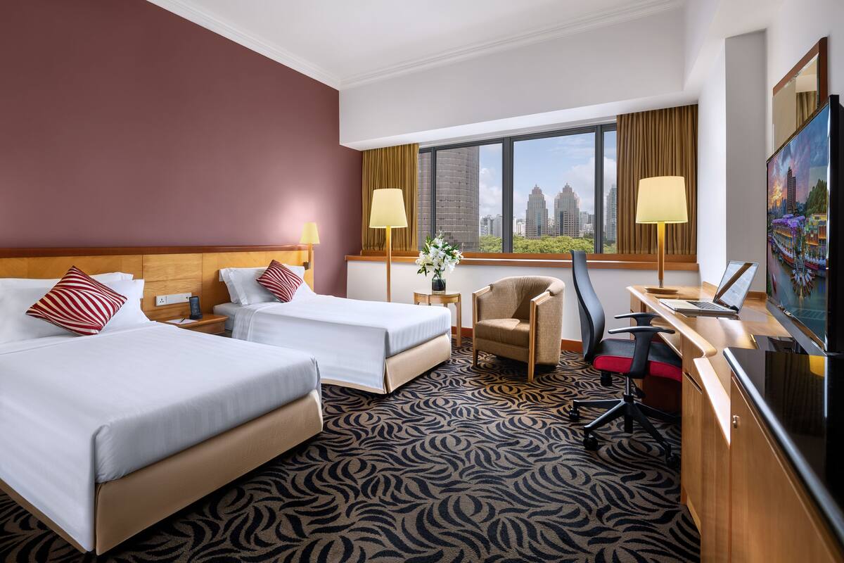 A hotel room with two beds, a desk with a laptop, a chair, a TV, and a large window with a city view.
