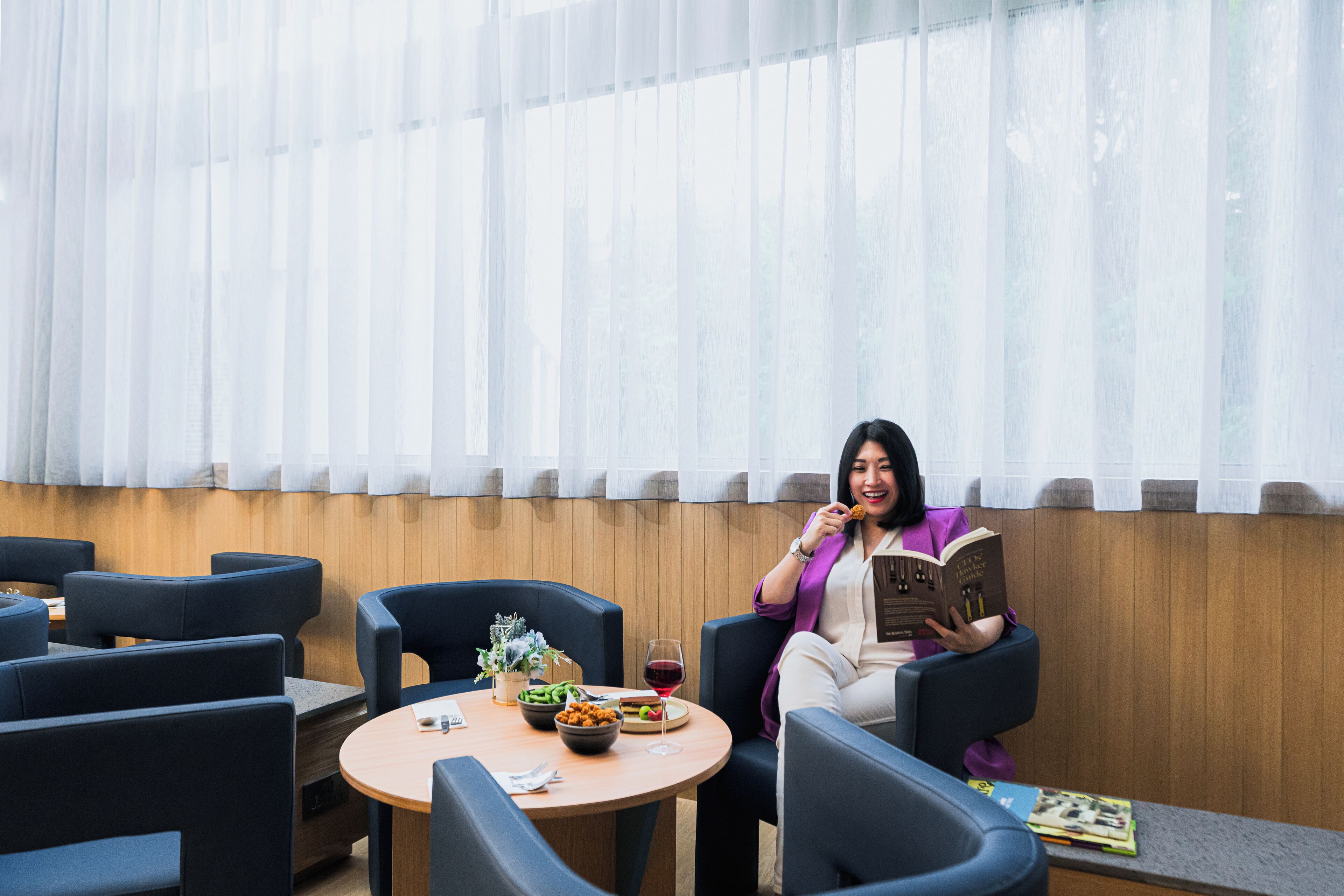 Una mujer sentada a una mesa redonda con un libro, una copa de vino tinto y un cuenco de fruta. La habitación tiene sillas azules y una pared de madera.