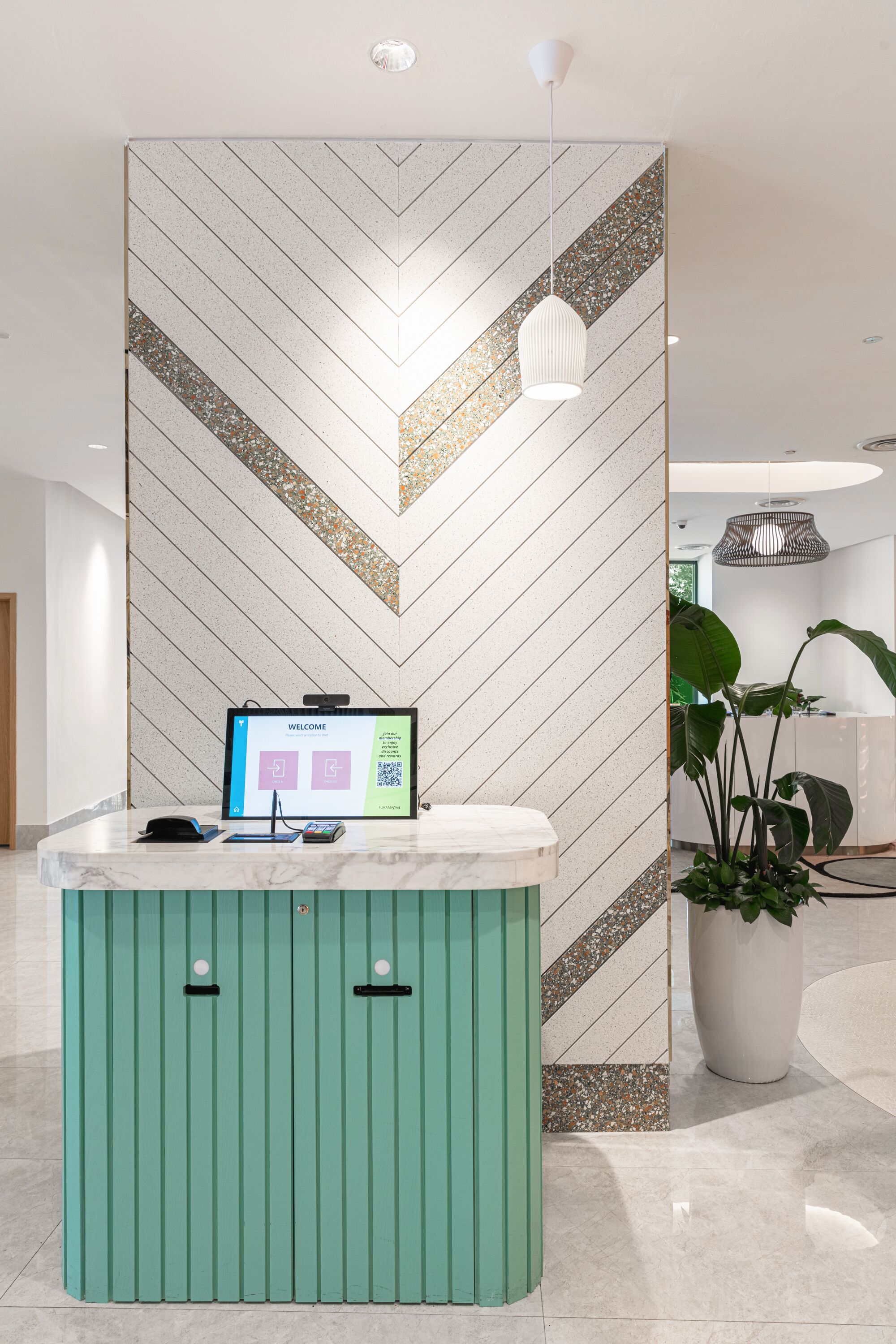 Un mostrador de recepción con una pantalla digital que muestra un mensaje de bienvenida, una planta en maceta y una pared con diseño en zigzag.
