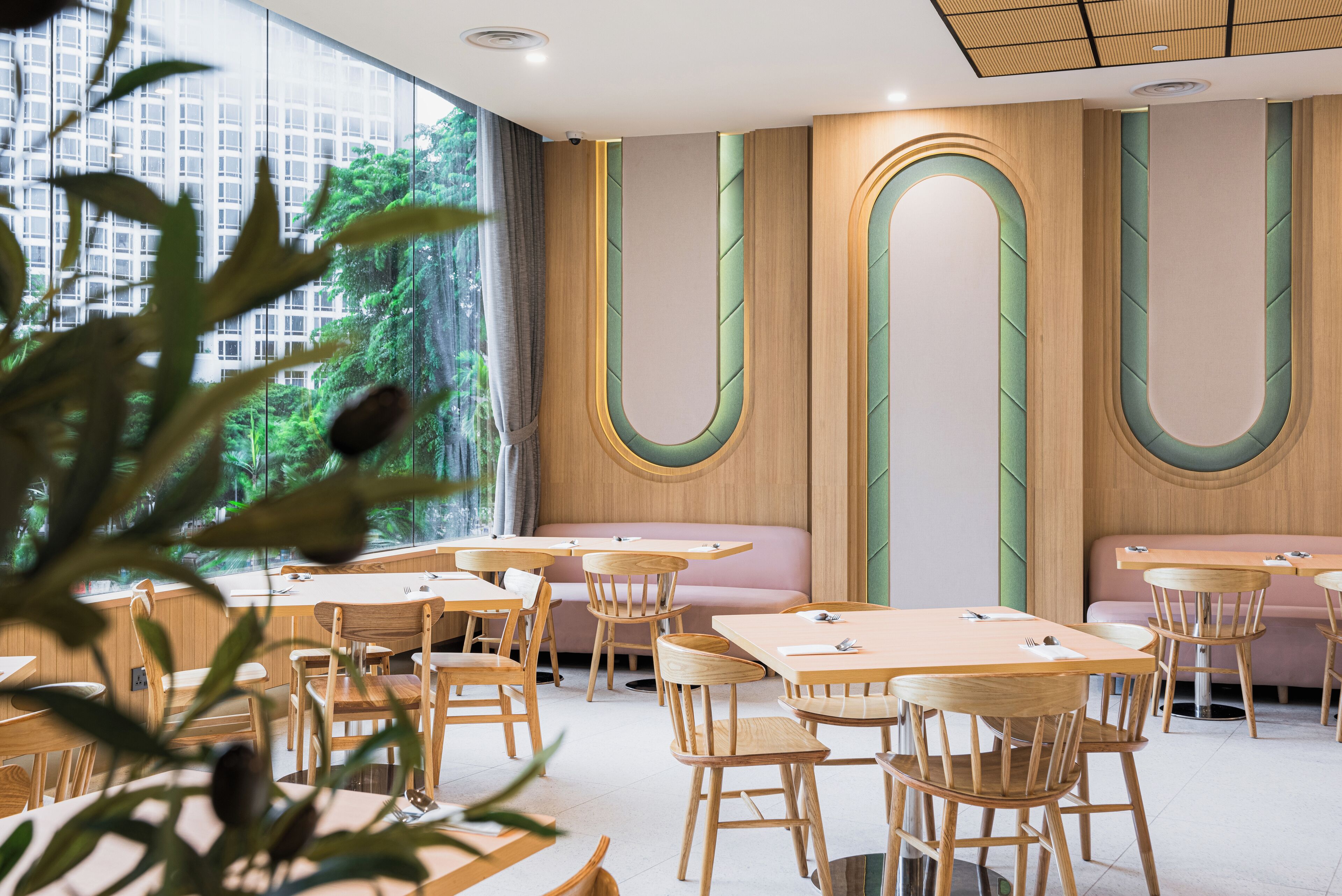 Interior moderno de restaurante con mesas y sillas de madera, asientos con cojines verdes y amplias ventanas que dan a zonas verdes.