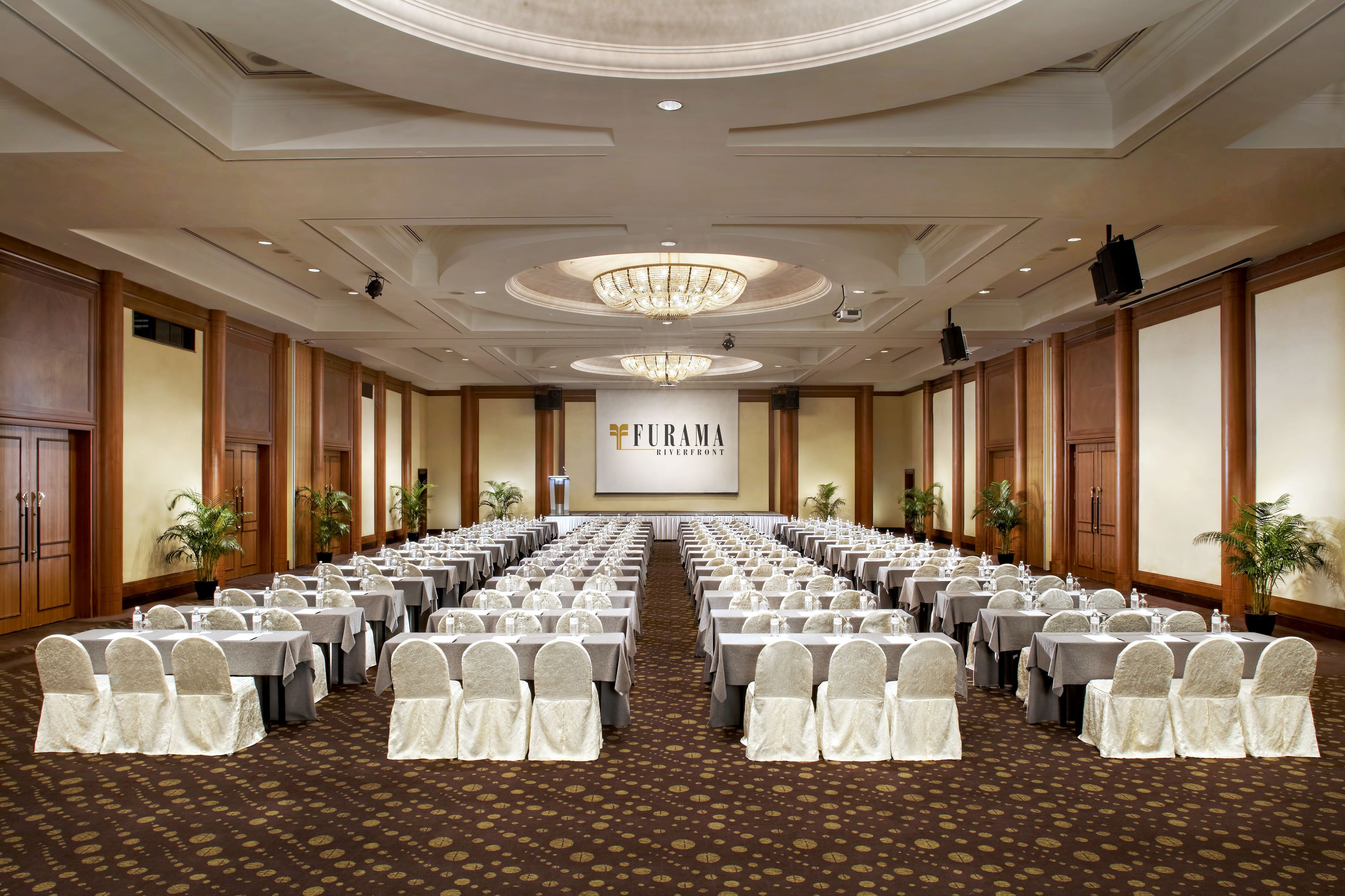 A conference room with rows of tables and chairs, a chandelier, and a banner reading "Furama Riverfront".