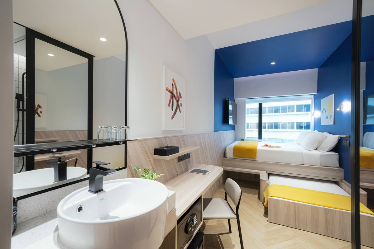 A modern hotel room with a large bathroom sink, a bedside table, a chair, and a bed with a yellow blanket.