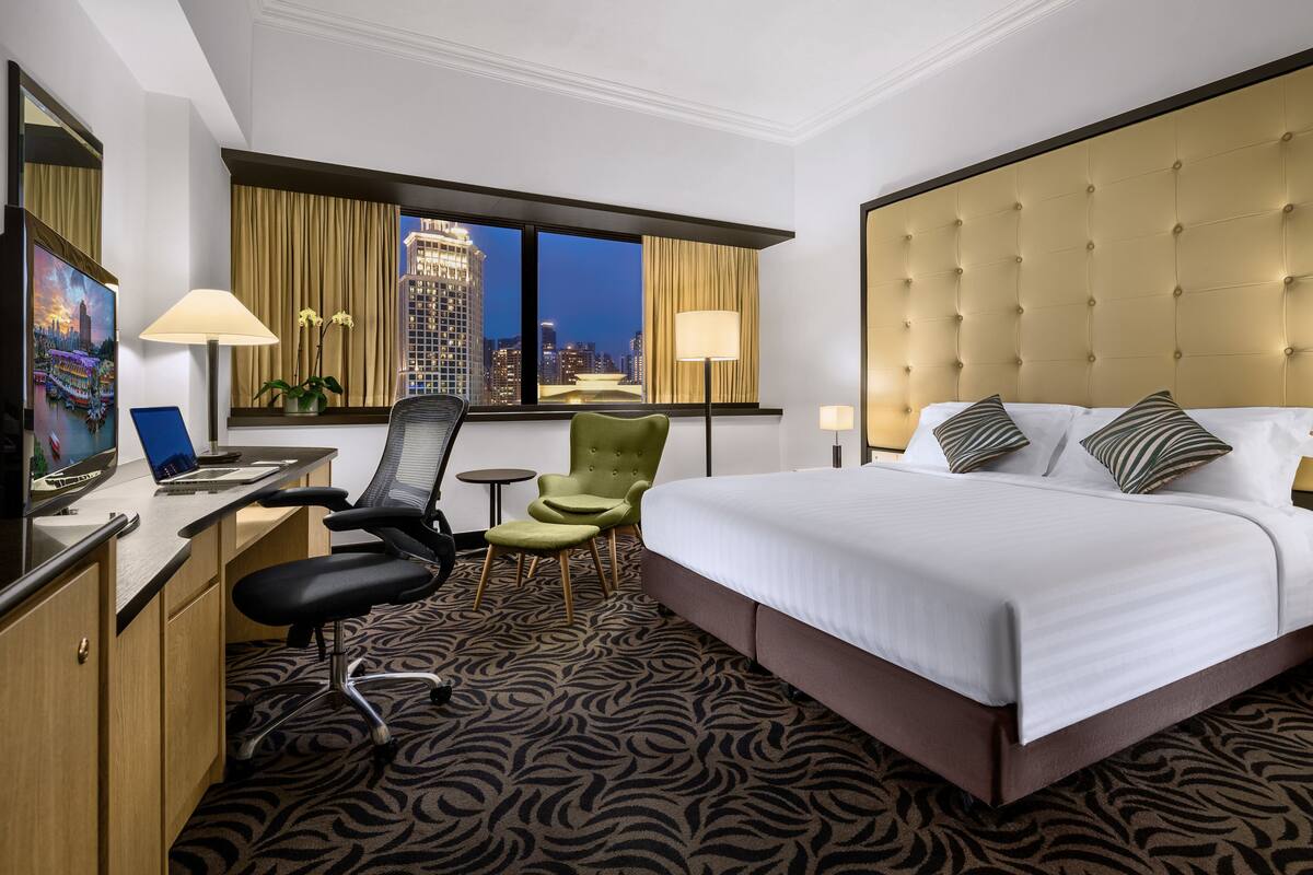 A hotel room with a bed, desk, chair, and a large window with a city view.