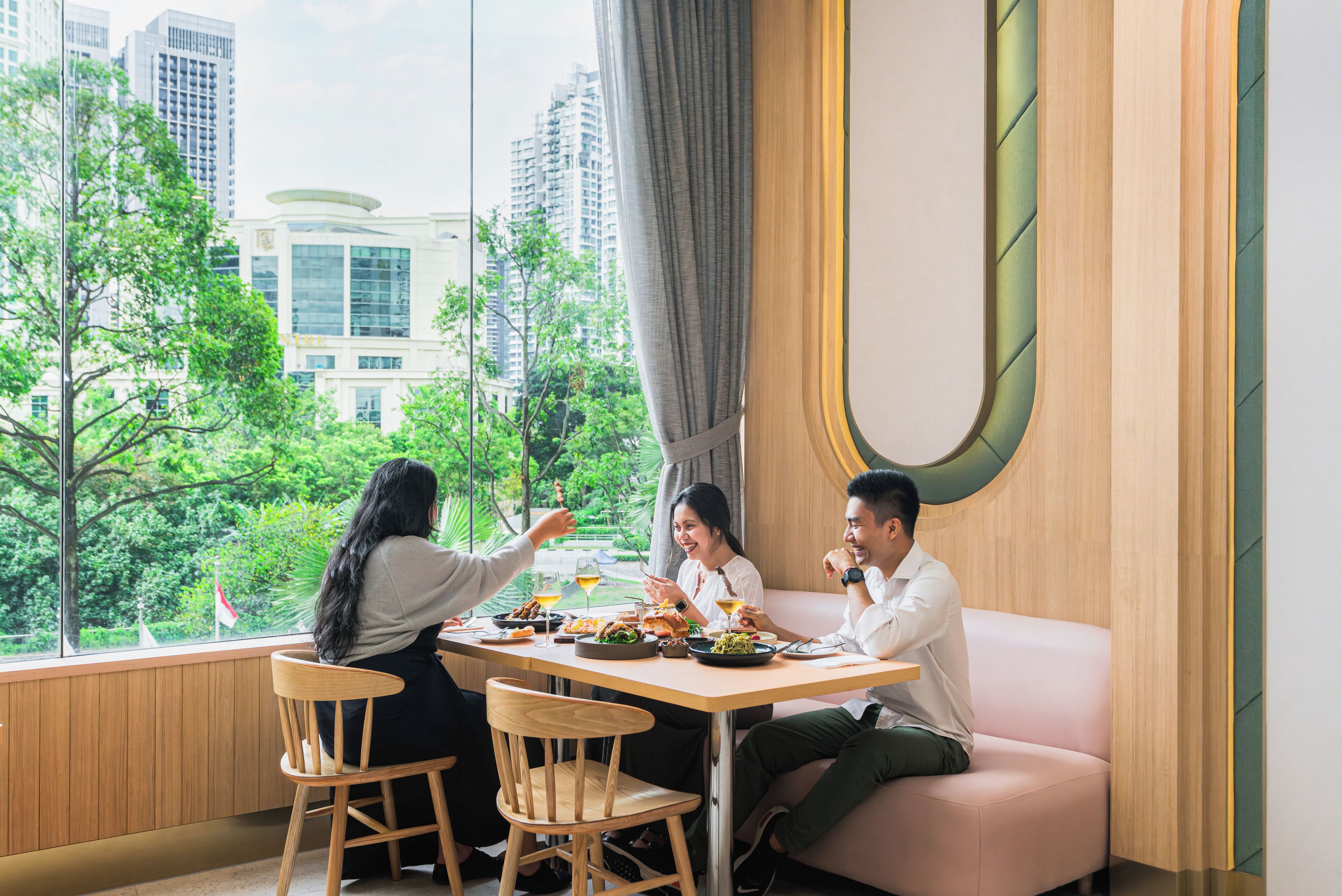 A dining area with a table set for three people, a pink sofa, and a view of greenery and buildings outside.