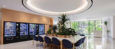 A modern dining area with a central table surrounded by chairs, a large plant, and vending machines in the background.