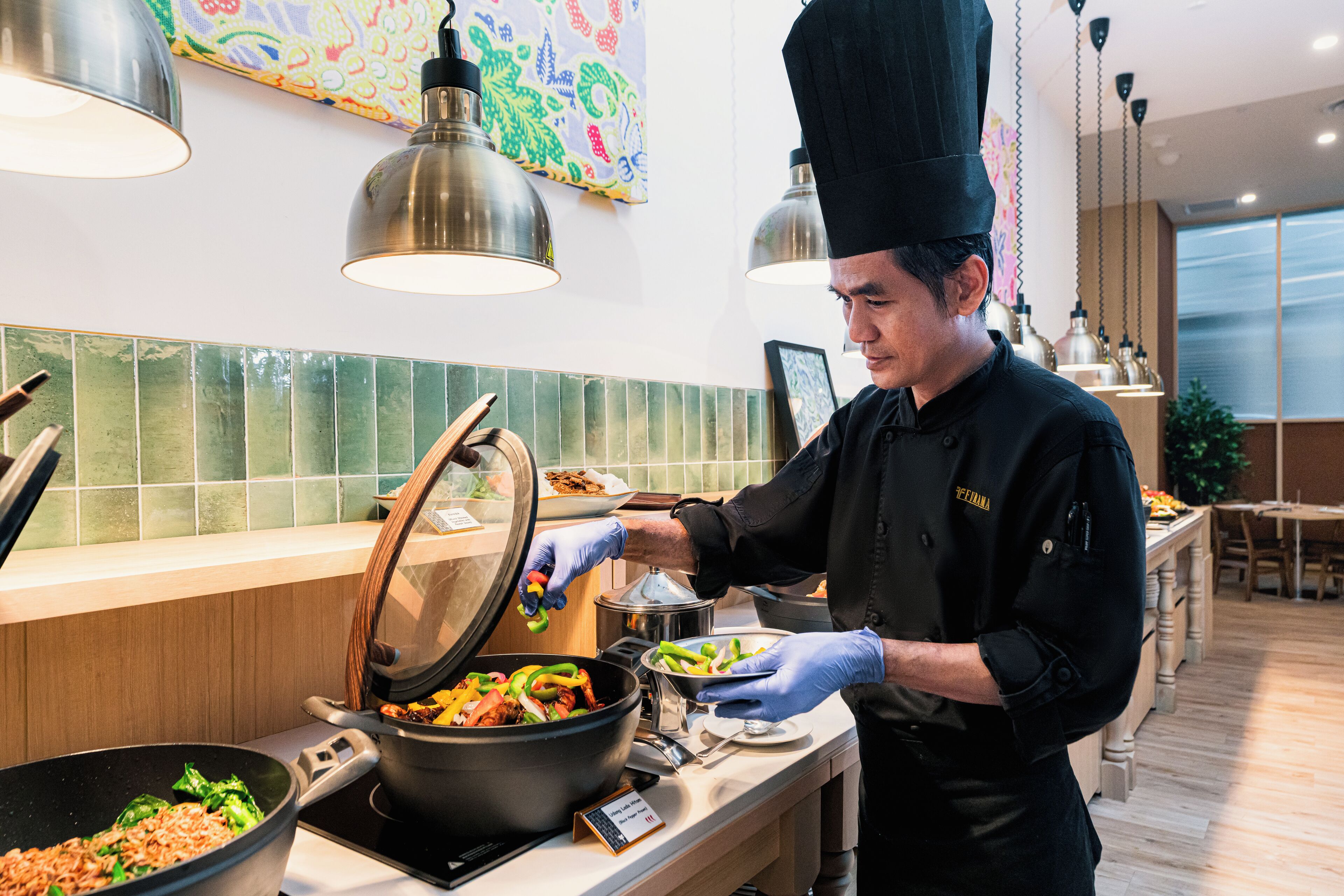 Een buffetopstelling met een kok die eten serveert, een grote pan met eten en een kleurrijke wanddecoratie.