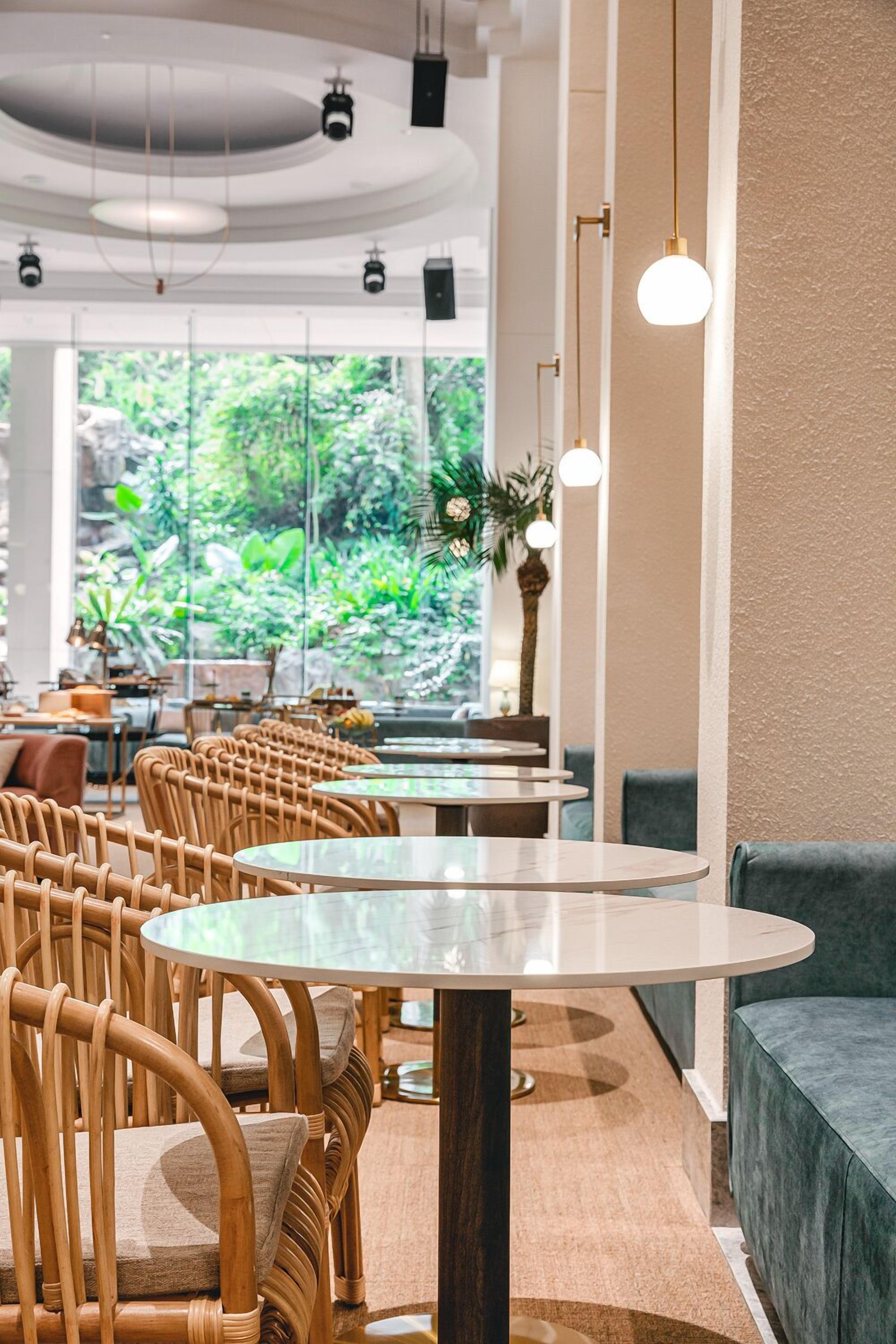 A restaurant interior with round tables, wicker chairs, and a view of greenery outside.