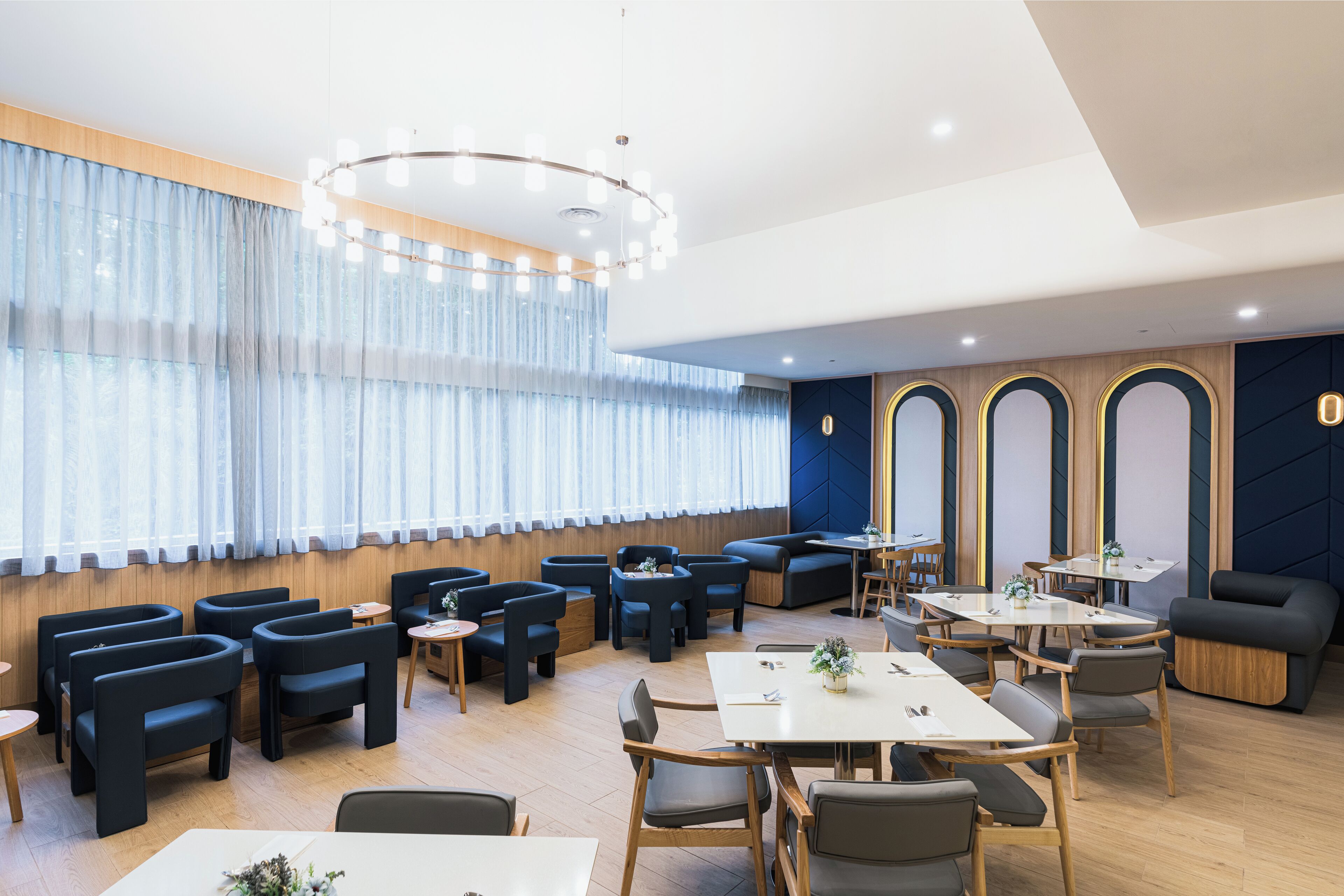 A modern dining area with round tables and black chairs, wooden accents, and a large window with sheer curtains.