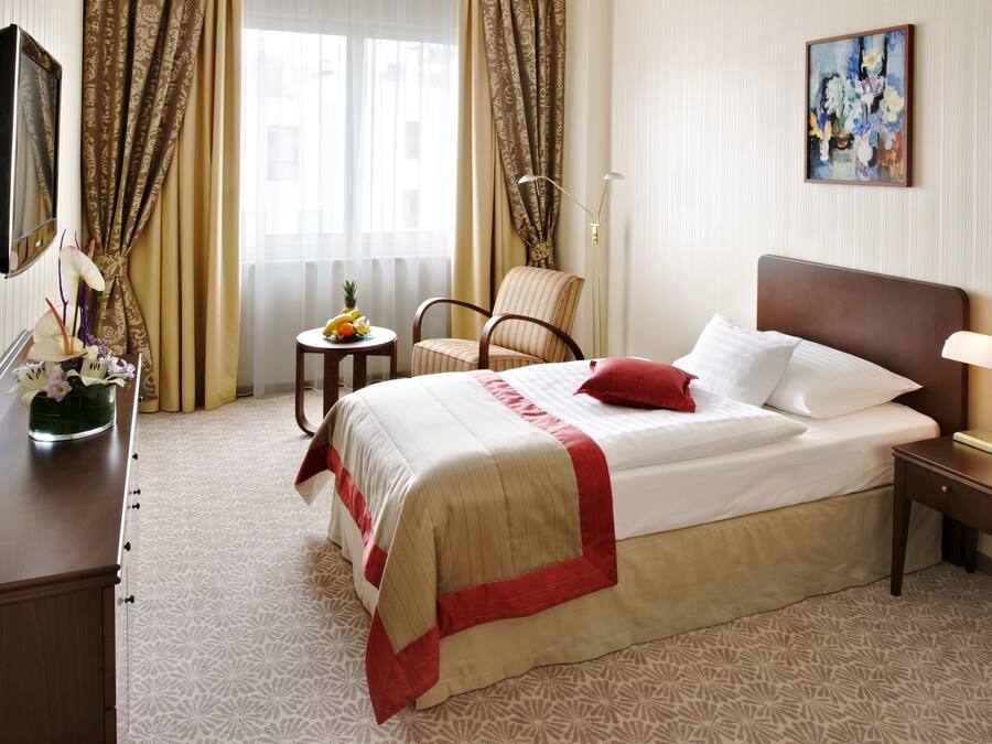 Une chambre d’hôtel avec un lit, une chaise, une petite table avec des fruits, un vase de fleurs et un tableau au mur.