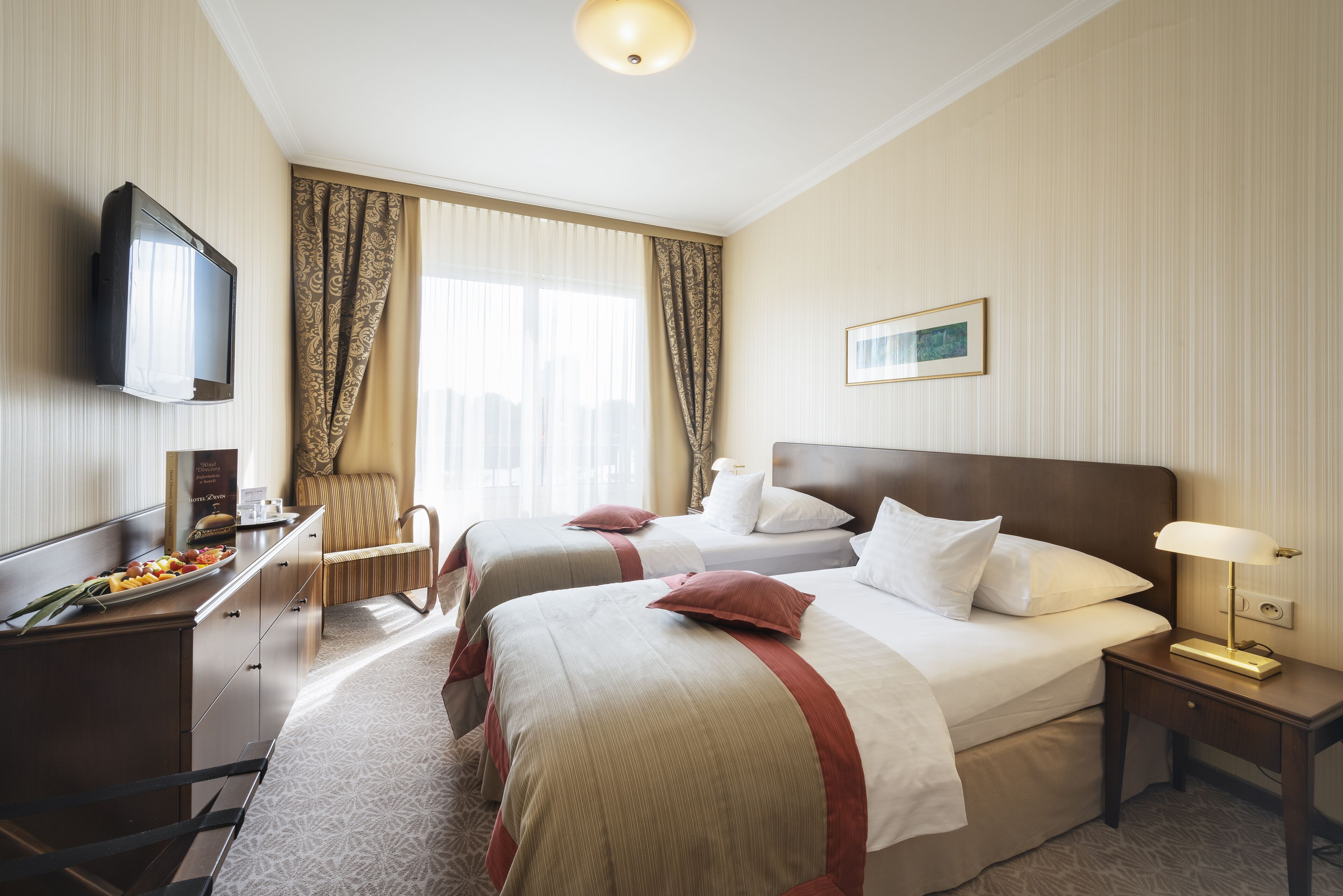 A hotel room with two beds, a desk with a TV, a chair, and a bedside table with a lamp.