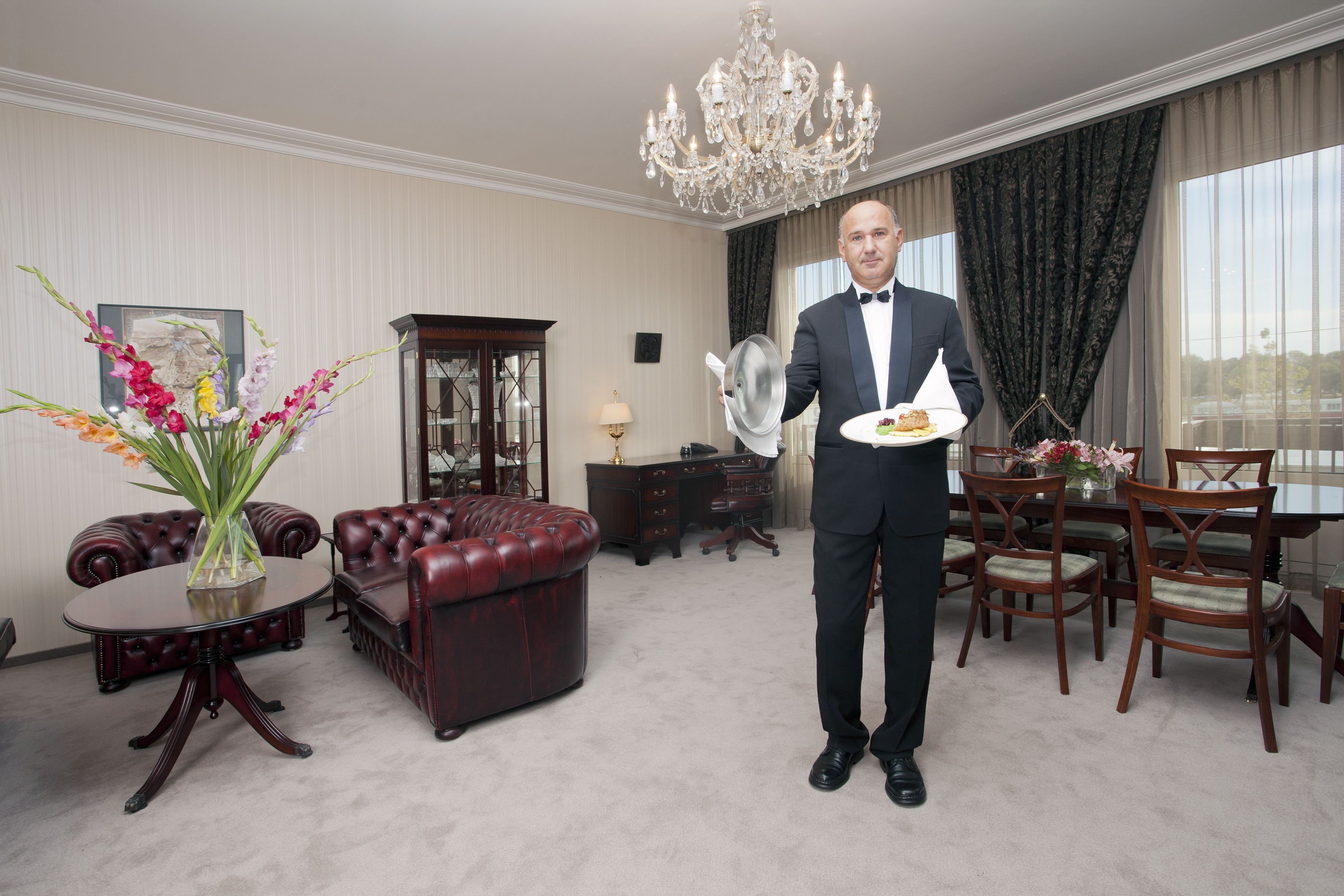A man in a formal suit holding a plate with food, standing in a room with a chandelier, leather furniture, and a large window.