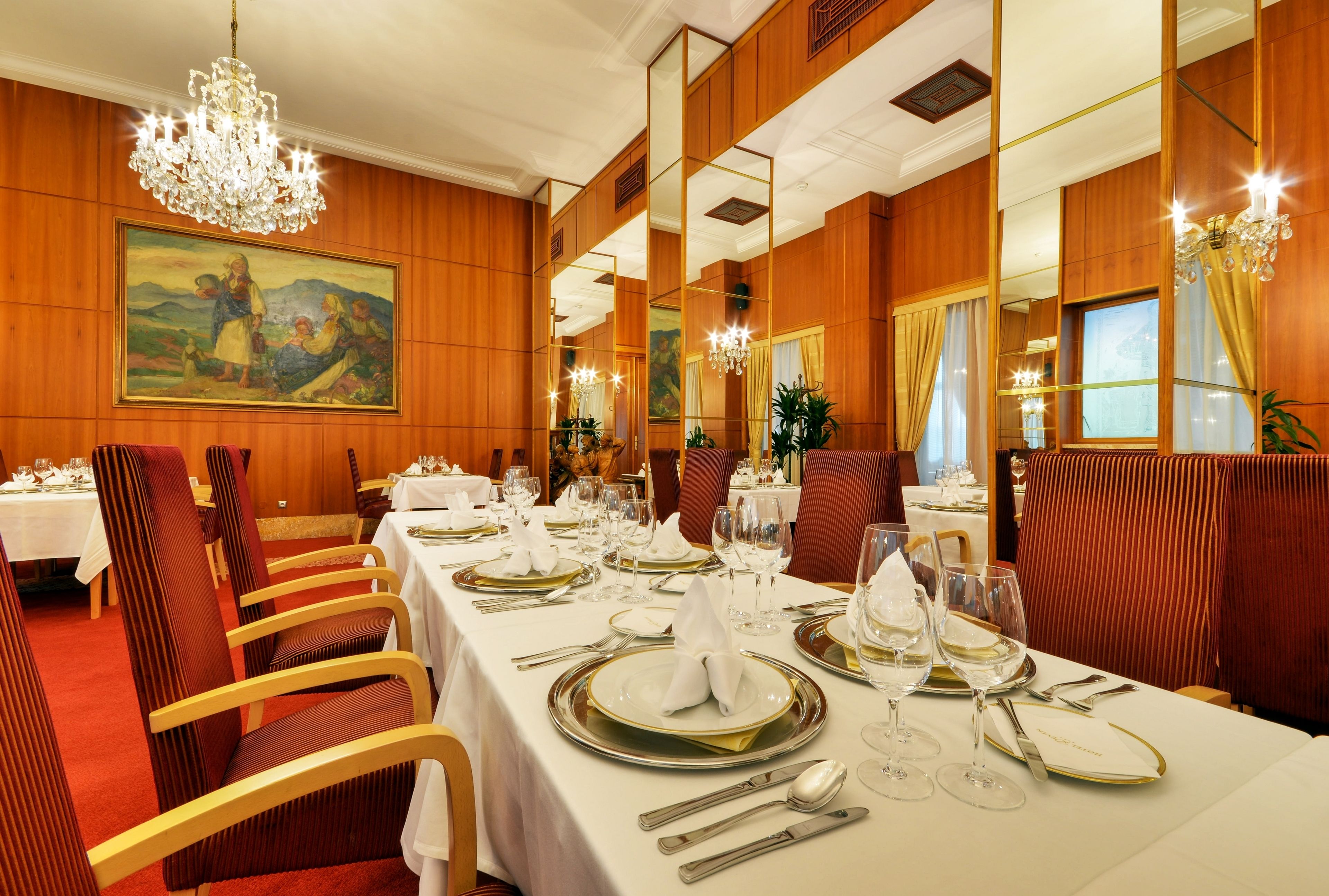 Une salle à manger aux murs lambrés, ornée d’un grand tableau et de lustres. Les tables sont dressées avec des nappes blanches, de l’argenterie et des assiettes dorées.