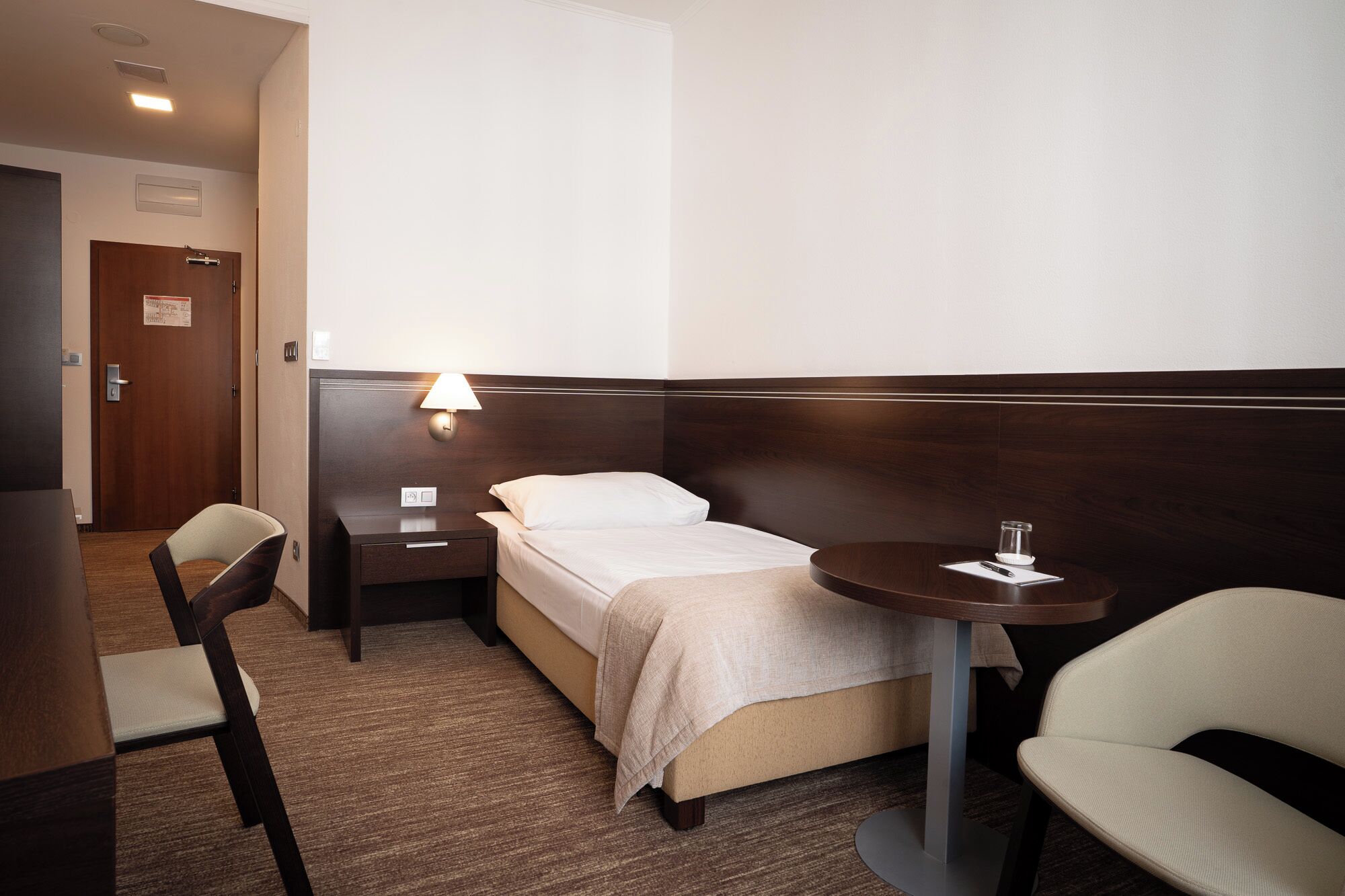 Et hotelværelse med seng, sengebord, stol og et lille bord med et glas.