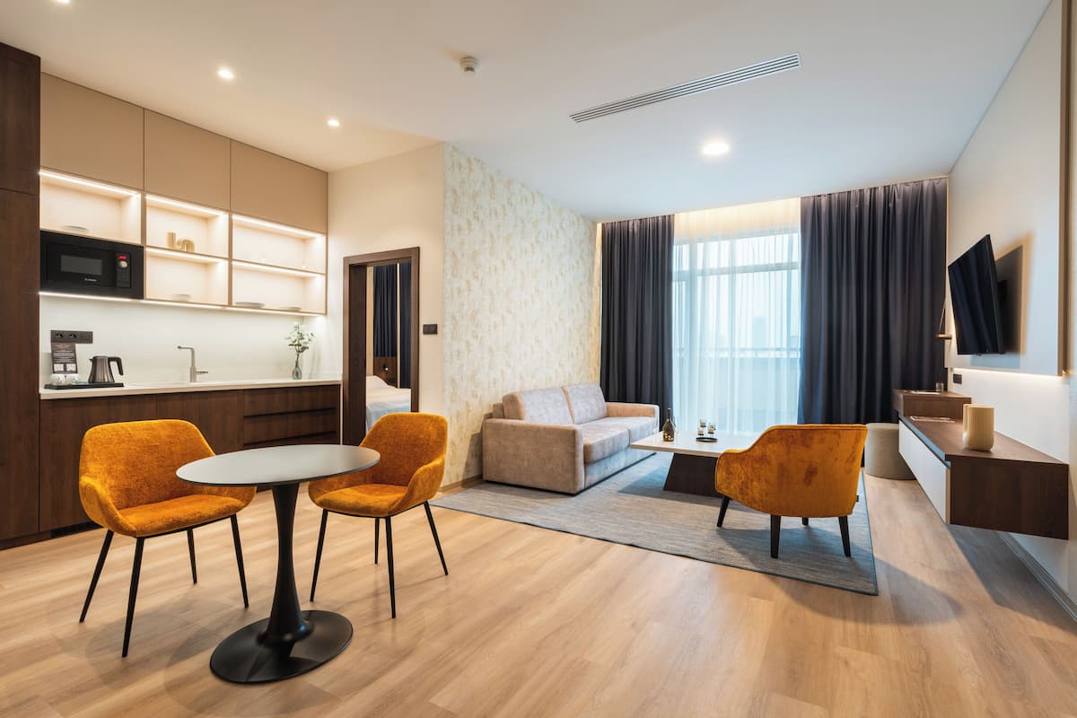 A modern hotel room with a kitchen, a dining area with a round table and two chairs, a sofa, and a TV mounted on the wall.