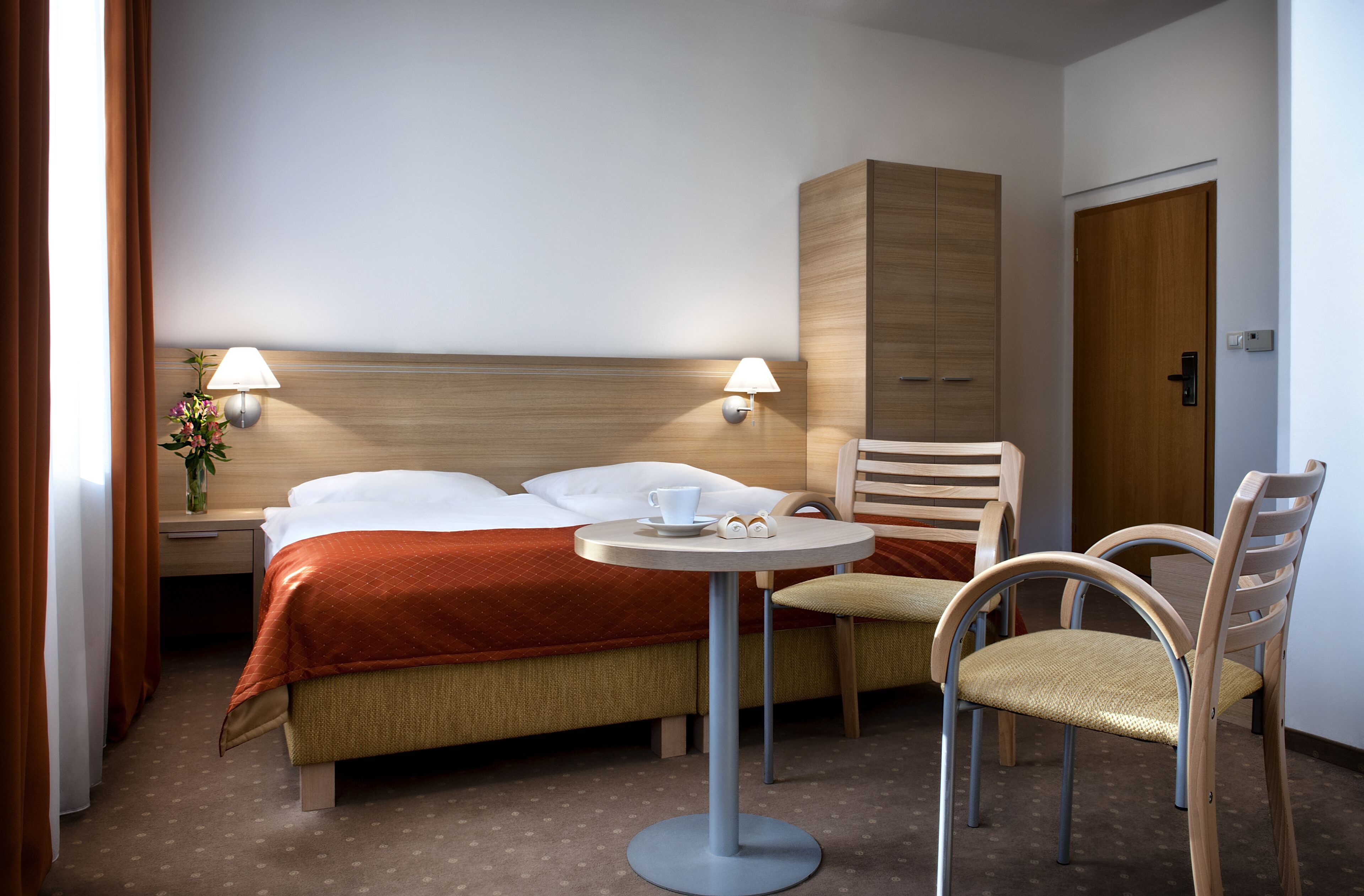 A hotel room with a bed, two bedside tables, a round table with a cup and saucer, two chairs, and a wooden wardrobe.