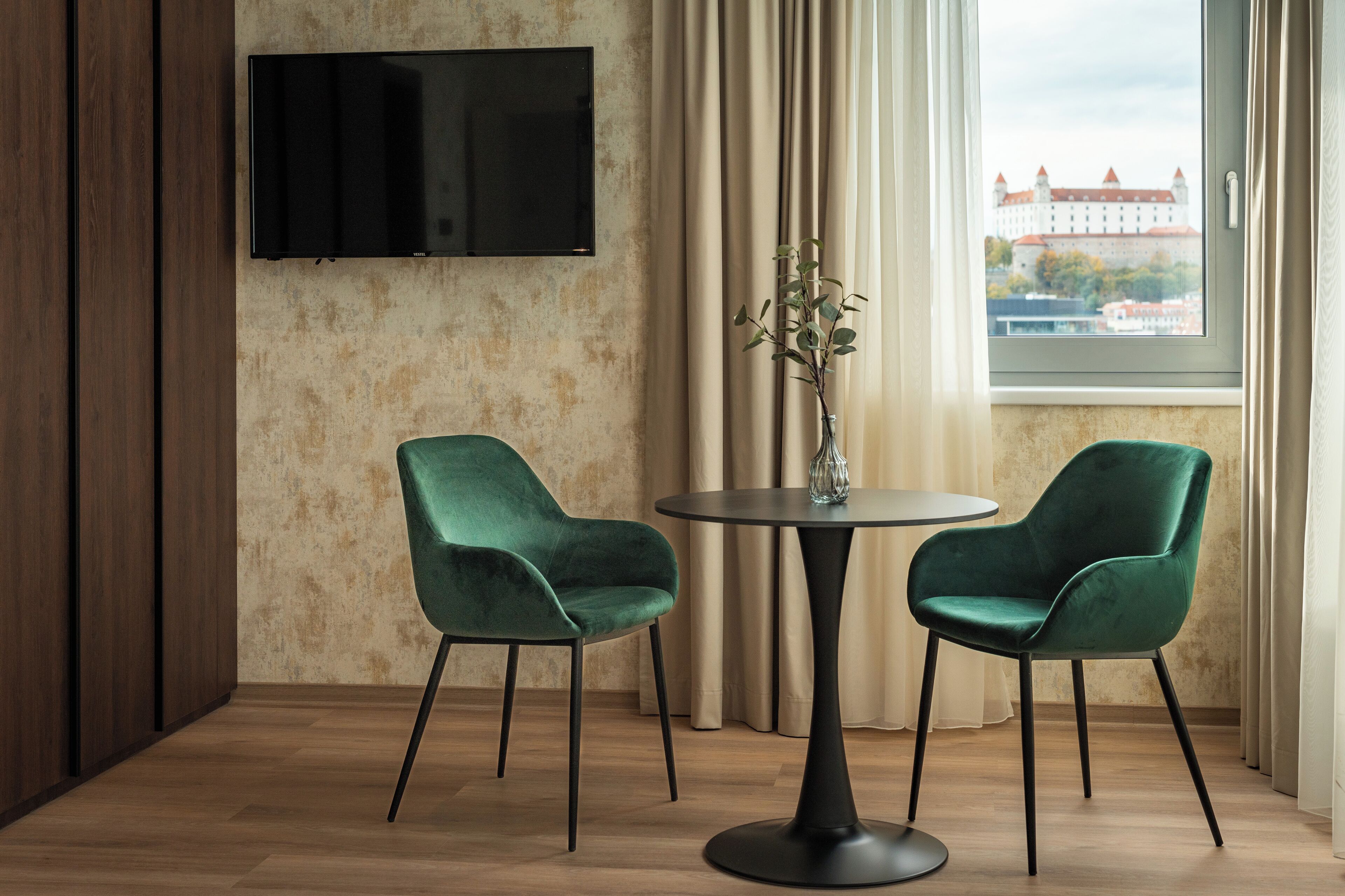 Et moderne værelse med to grønne stole, et rundt bord, en vase med en plante, et fladskærms-tv på væggen og et vindue med udsigt til en bygning.